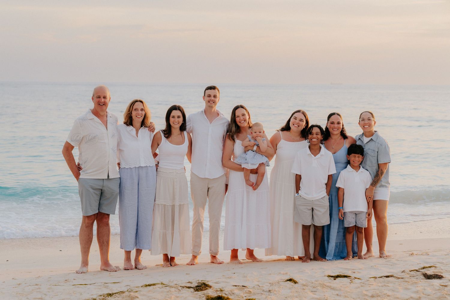 A group in coordinated white and beige outfits poses together on a sandy beach at sunset.