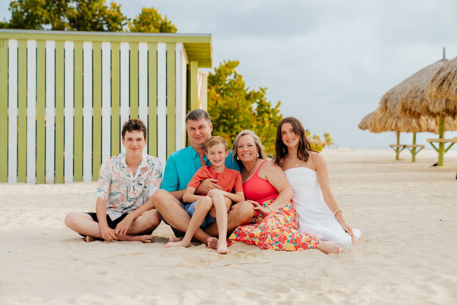 A family group sits together on a beach with a thatched hut and green wall in the background.