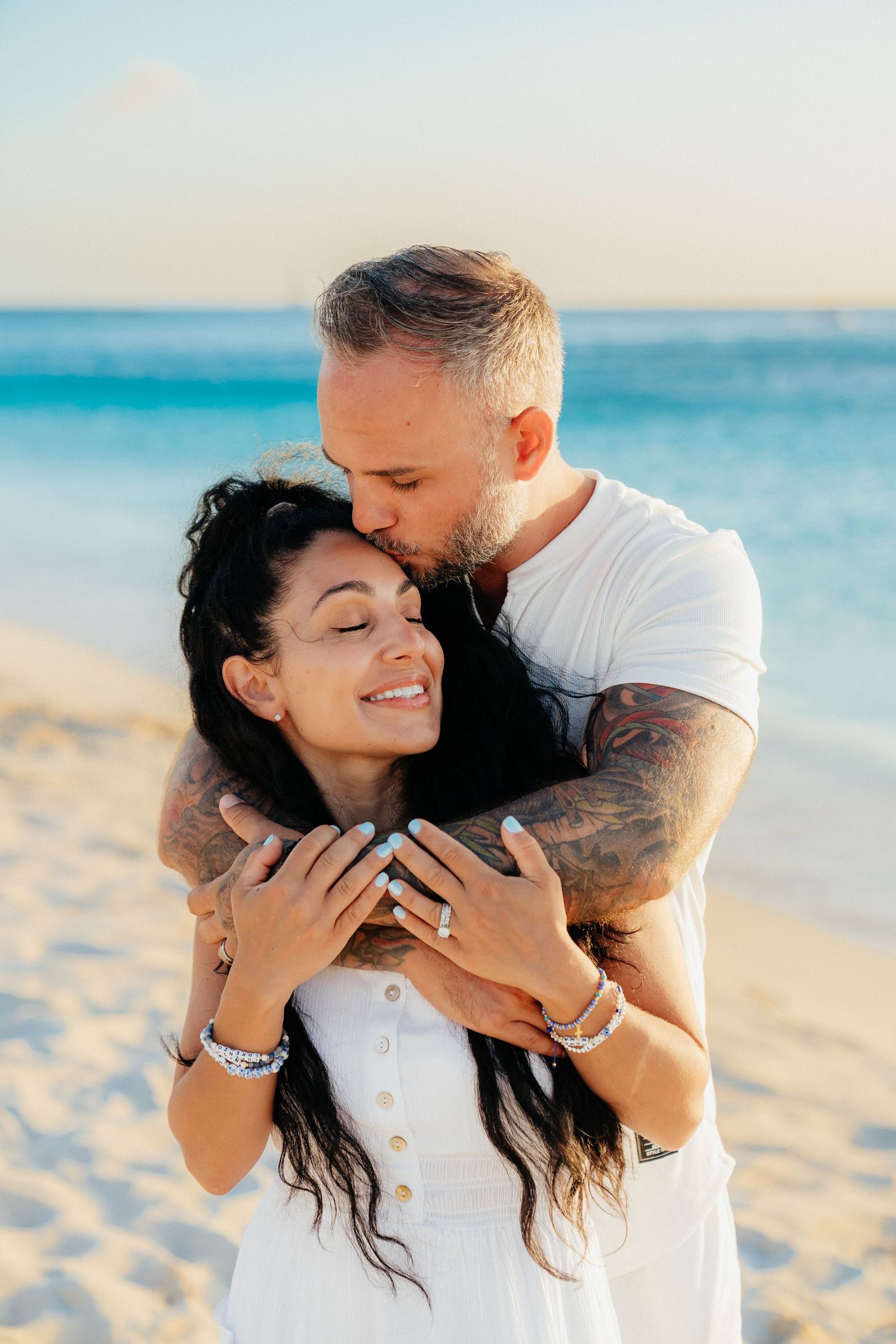 An intimate embrace captured on a sunny beach with crystal blue waters in the background.