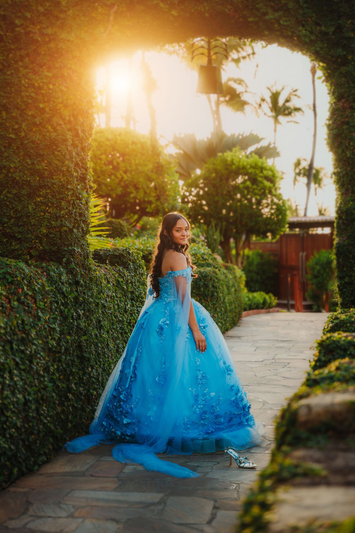 A flowing blue princess dress twirls in golden sunset light along a garden pathway.
