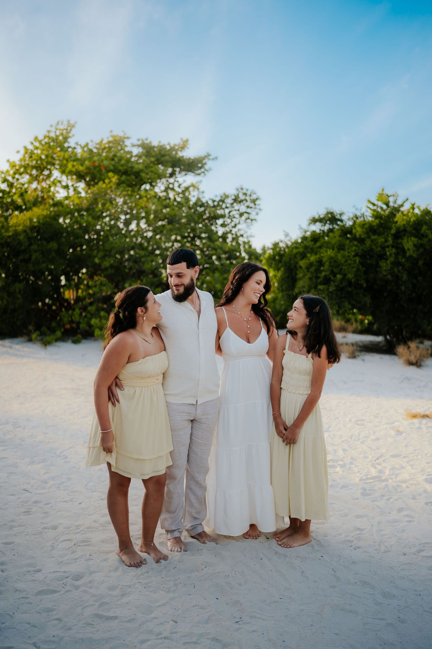 Family of four posing on white sand beach, couple in white wedding attire with two girls in yellow dresses.