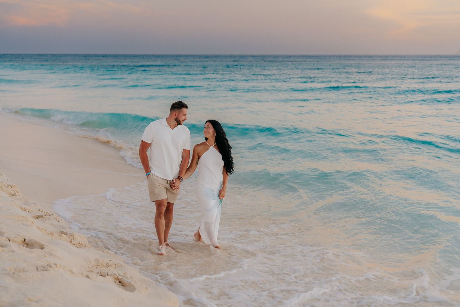 A couple walks hand in hand along a pristine beach at sunset.