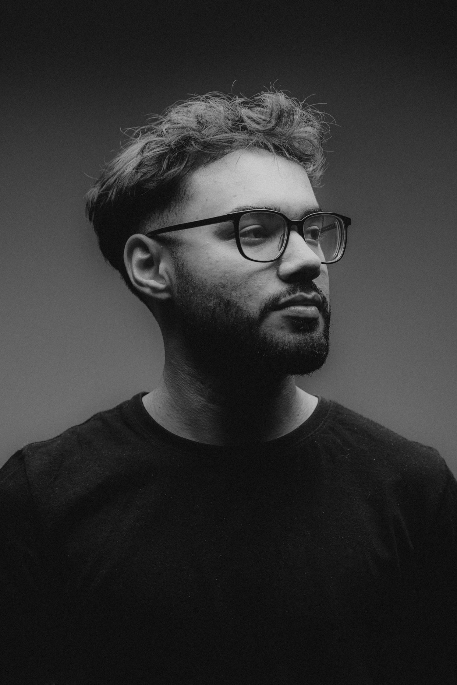 Black and white portrait of a person with glasses and a beard wearing a dark t-shirt against a simple background.
