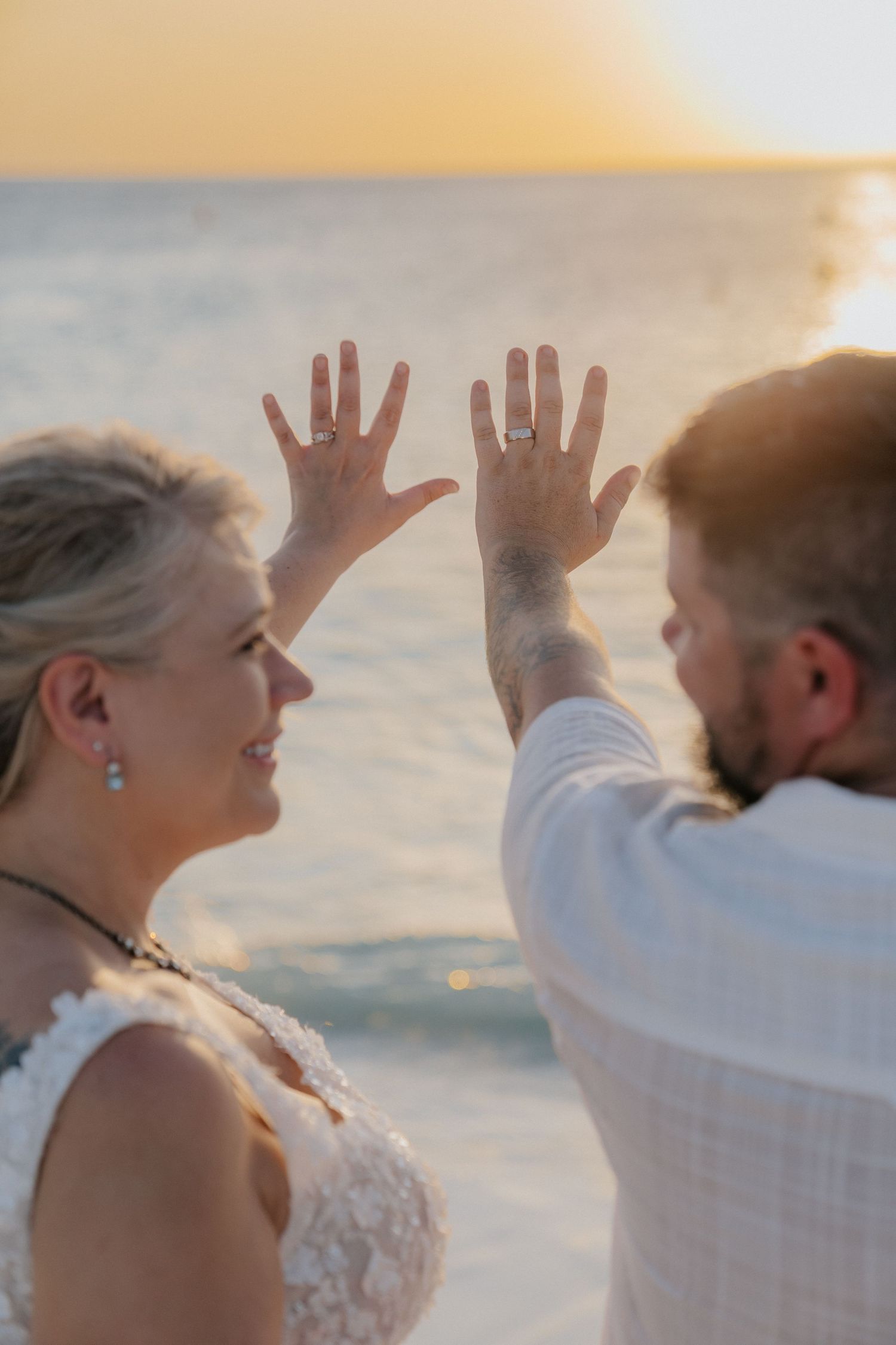 Couple showing their wedding rings against sunset ocean backdrop.