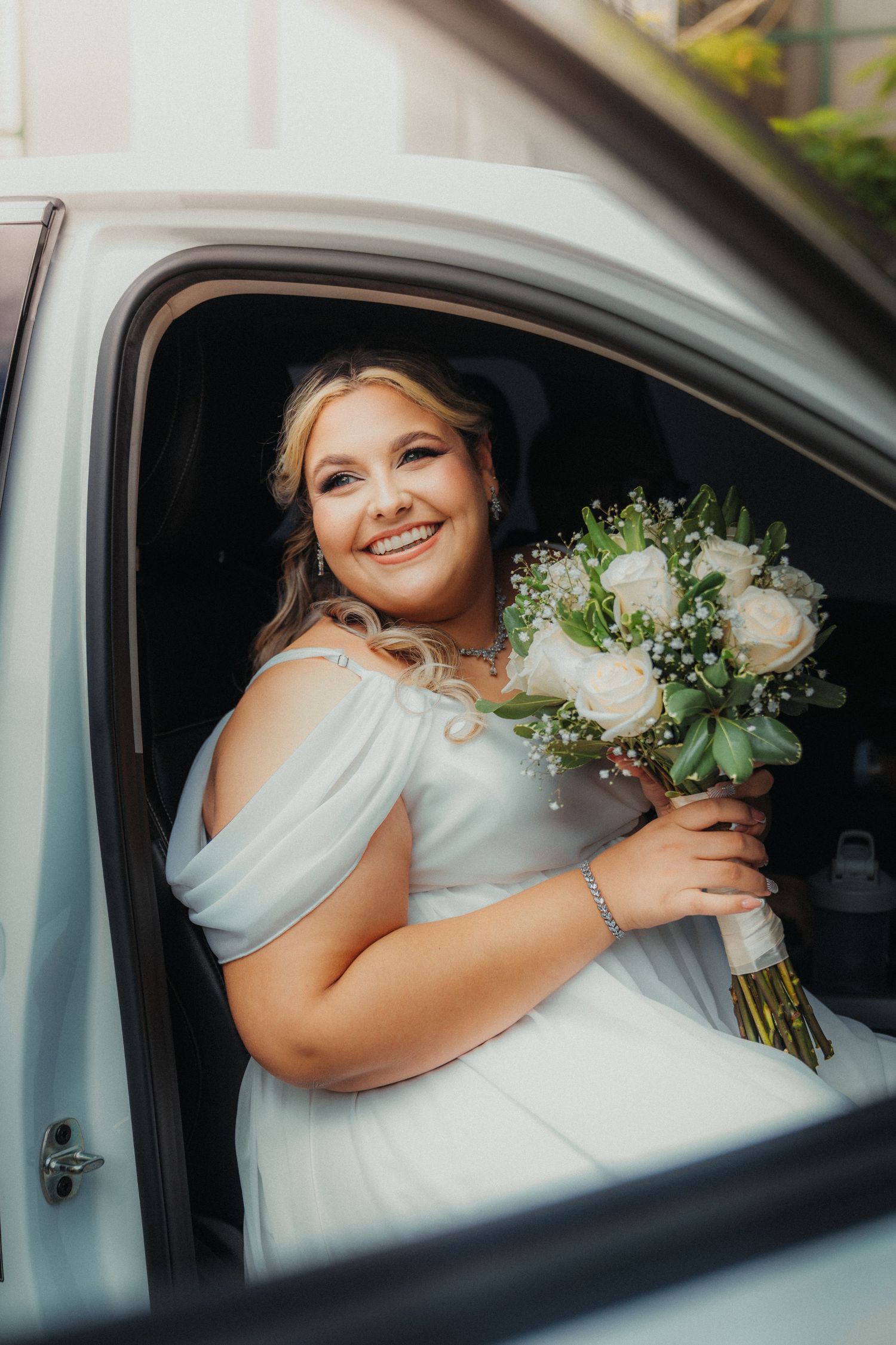 Person in white dress holds bouquet while sitting in car with joyful expression.