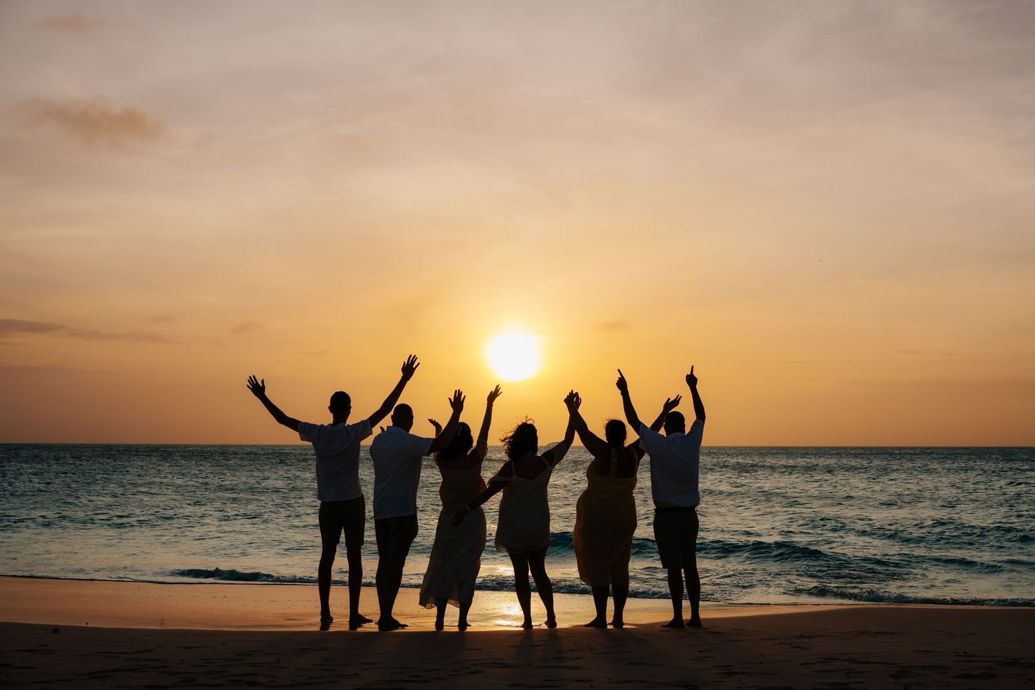 Silhouettes of six friends raising arms at sunset on a tropical beach with golden sky and ocean backdrop.