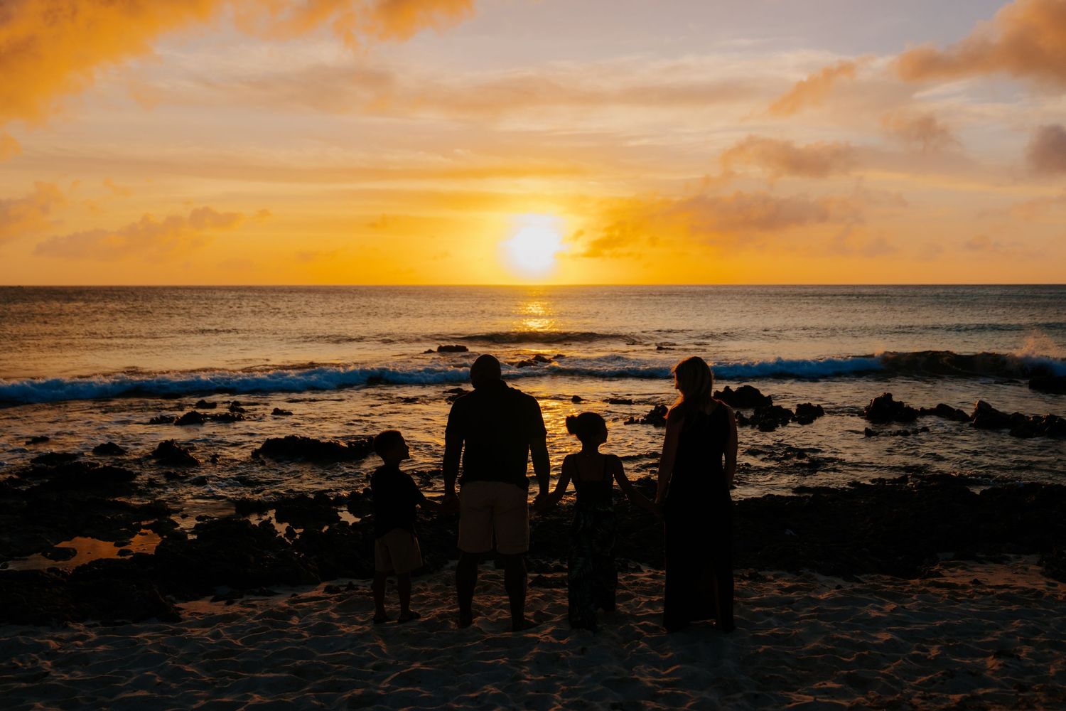 Silhouette of a family watching a stunning golden sunset over the ocean on a rocky beach.