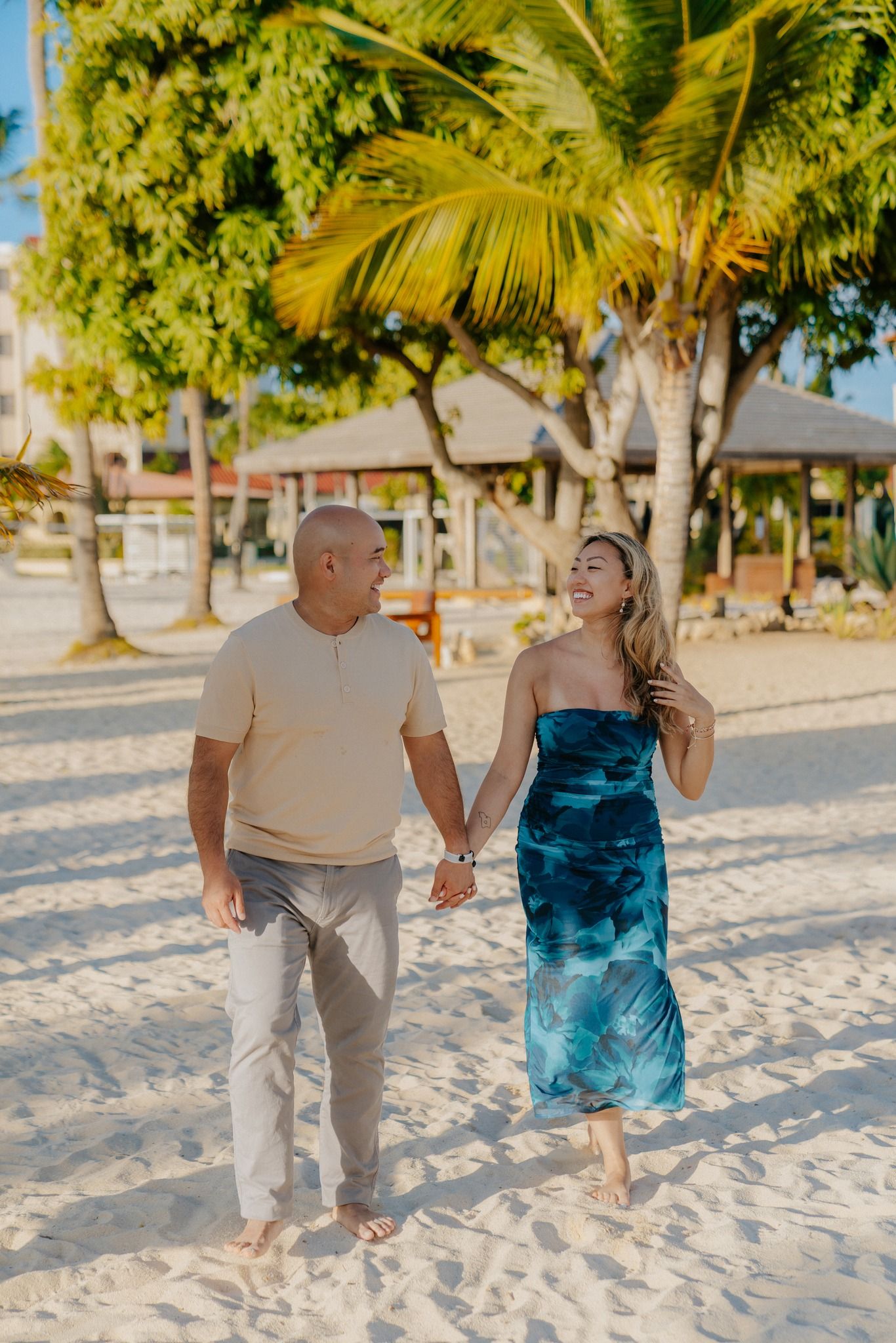 Couple sharing laughter while walking on sandy beach with palm trees and cabanas in background.