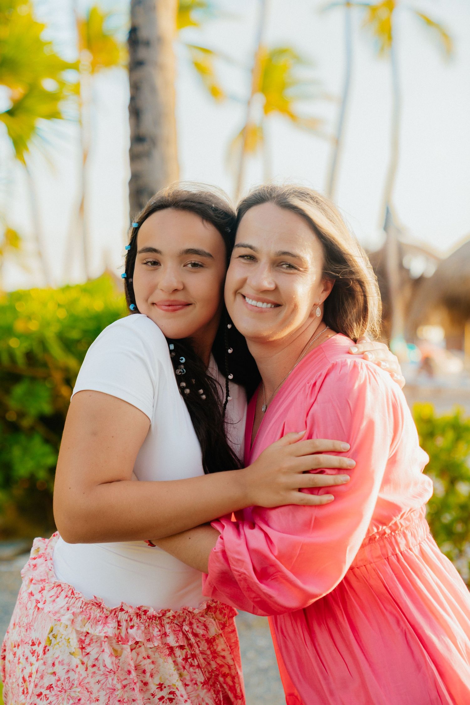 Two people share an affectionate embrace while wearing pink and white floral dresses among palm trees.