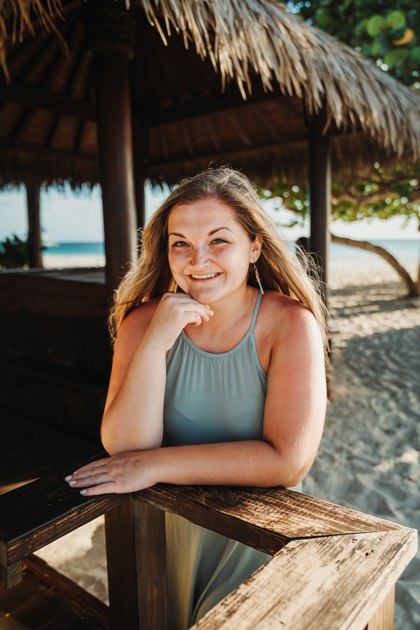 A person smiling while sitting at a wooden table under a thatched beach umbrella.