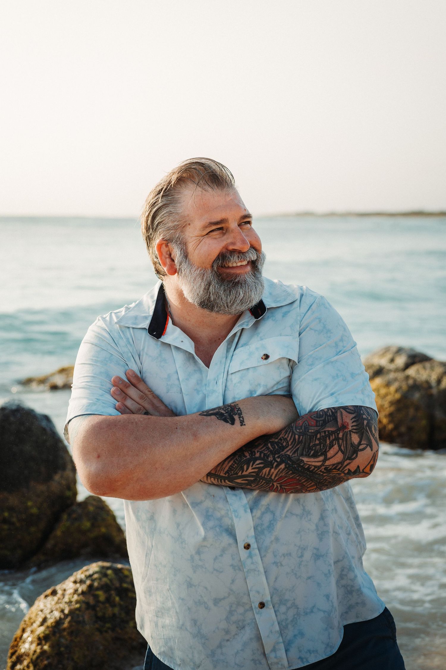 Man in light blue shirt smiling while sitting by coastal rocks at the ocean.
