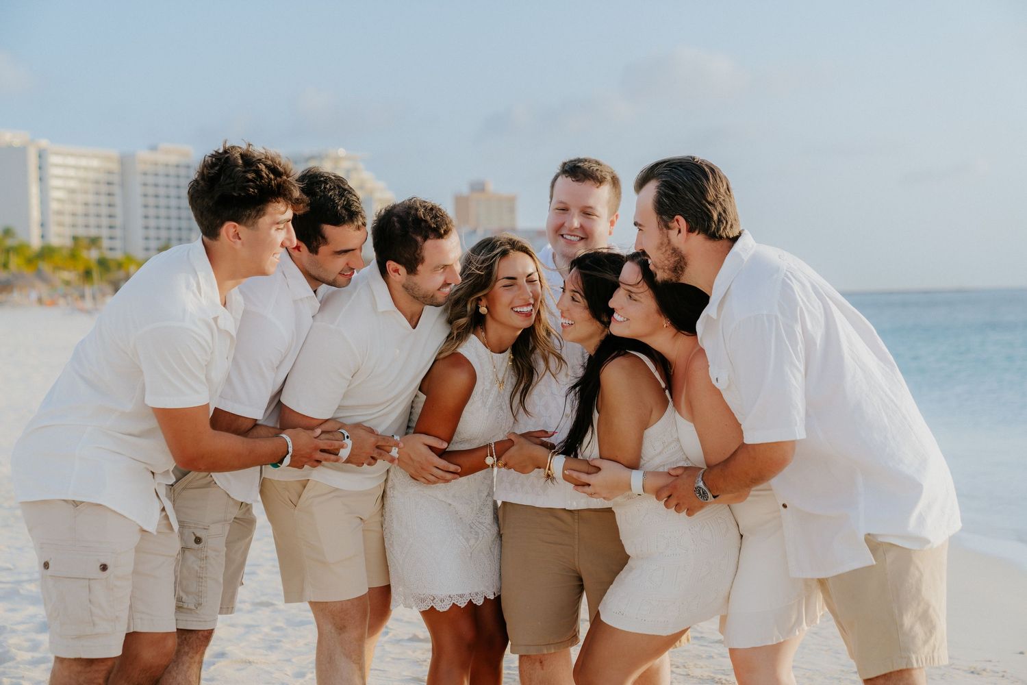 Group photo session on a white sand beach with everyone wearing coordinated light colored outfits.