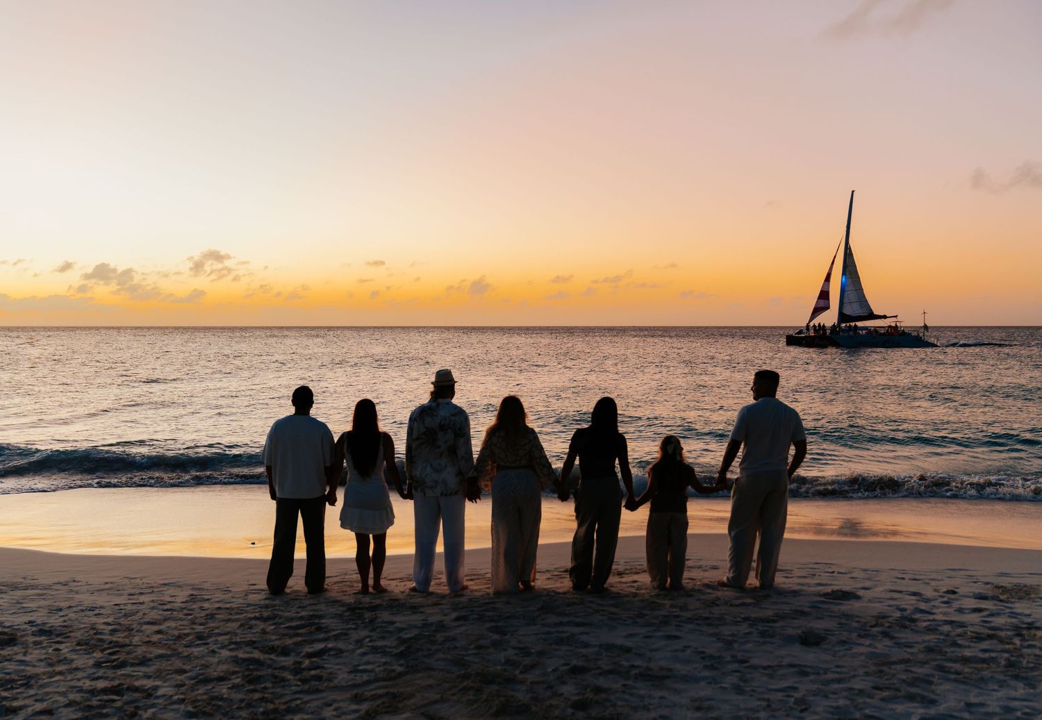Silhouettes of people enjoying a sunset on the beach with sailboats on the horizon.