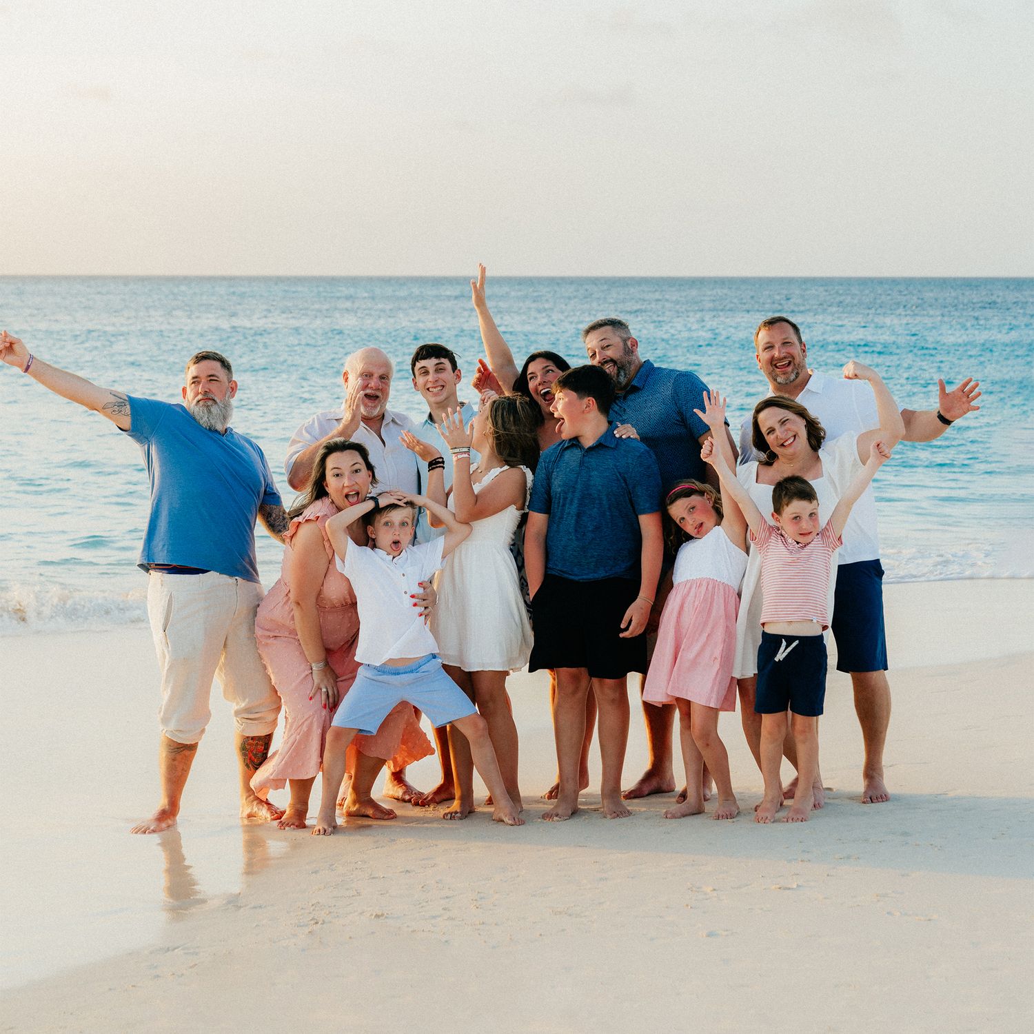 A large group poses playfully together on a white sand beach with turquoise water in the background.