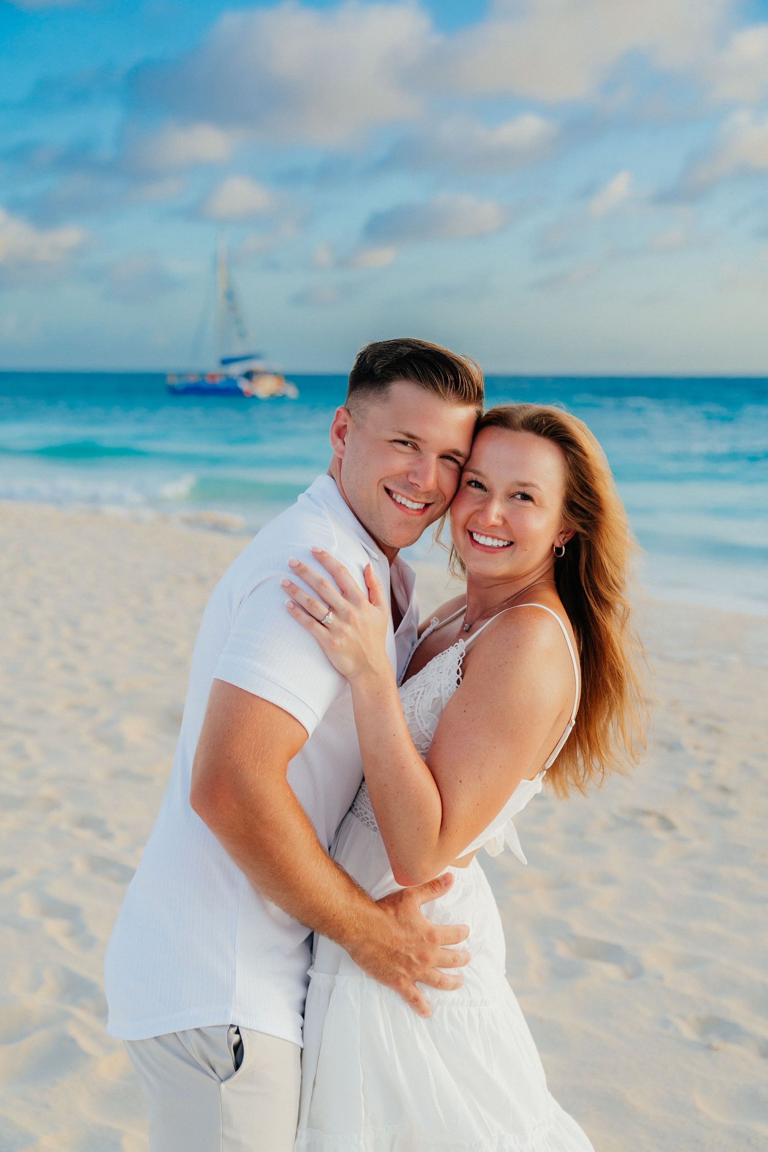 A romantic beach portrait with a sailboat visible in the turquoise waters behind.