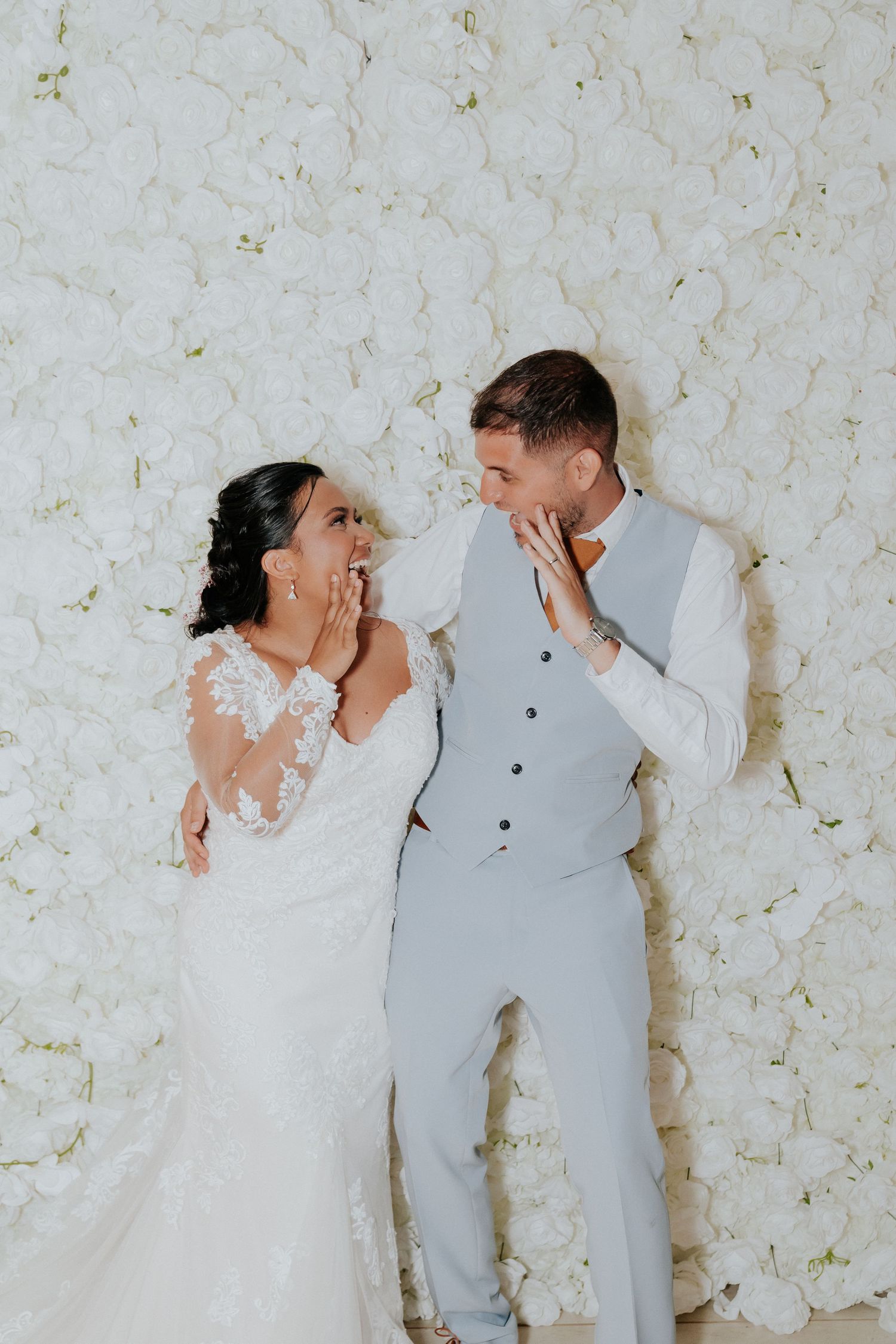 Wedding couple poses playfully against a white floral wall backdrop.