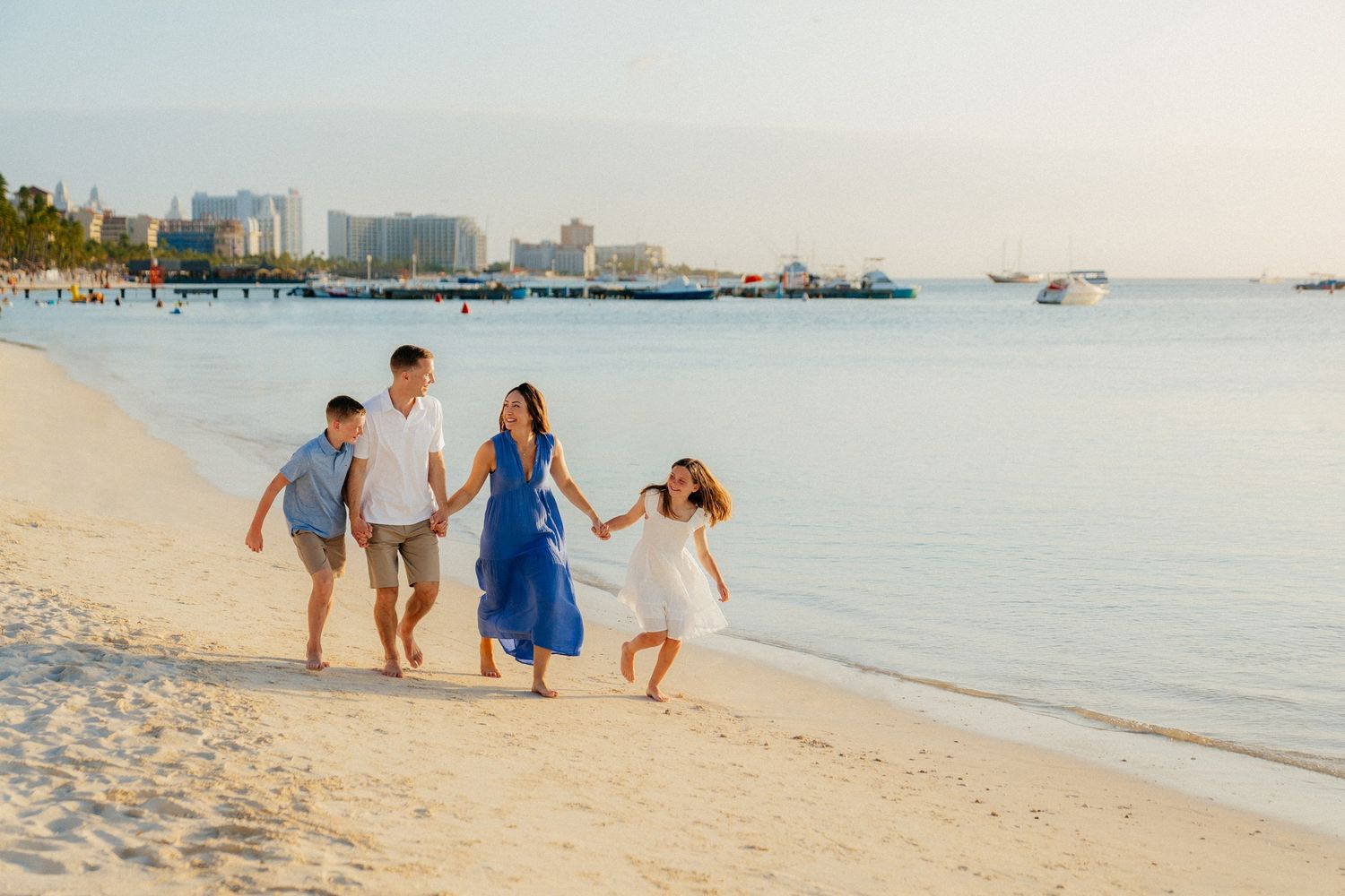 Family walking together along white sandy beach with city skyline and boats in background.