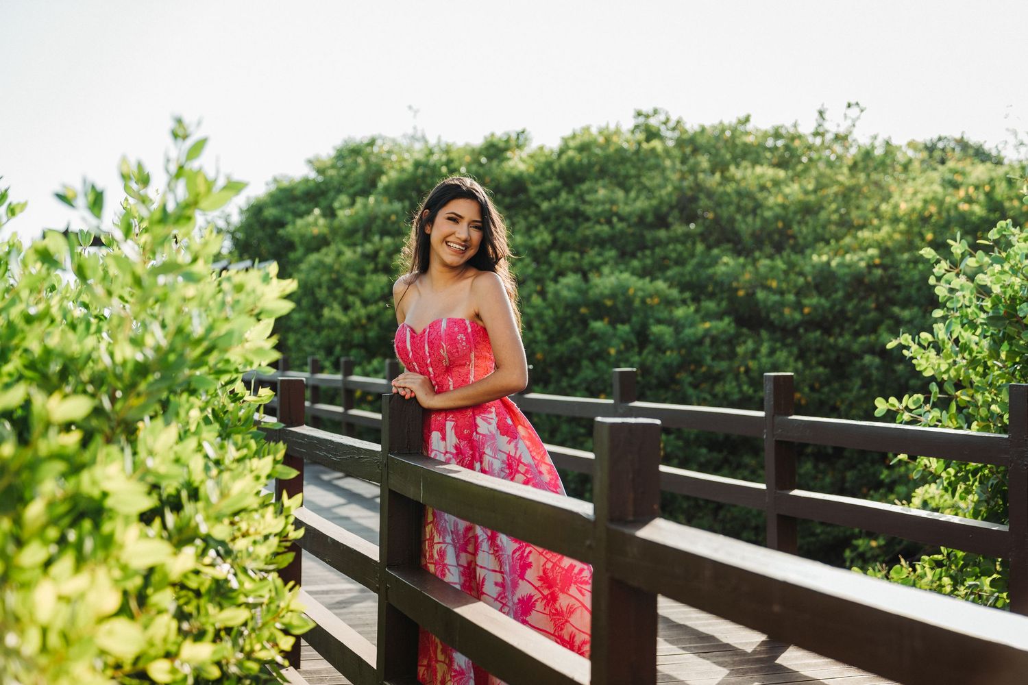 Woman in red floral dress standing on wooden bridge pathway surrounded by greenery.