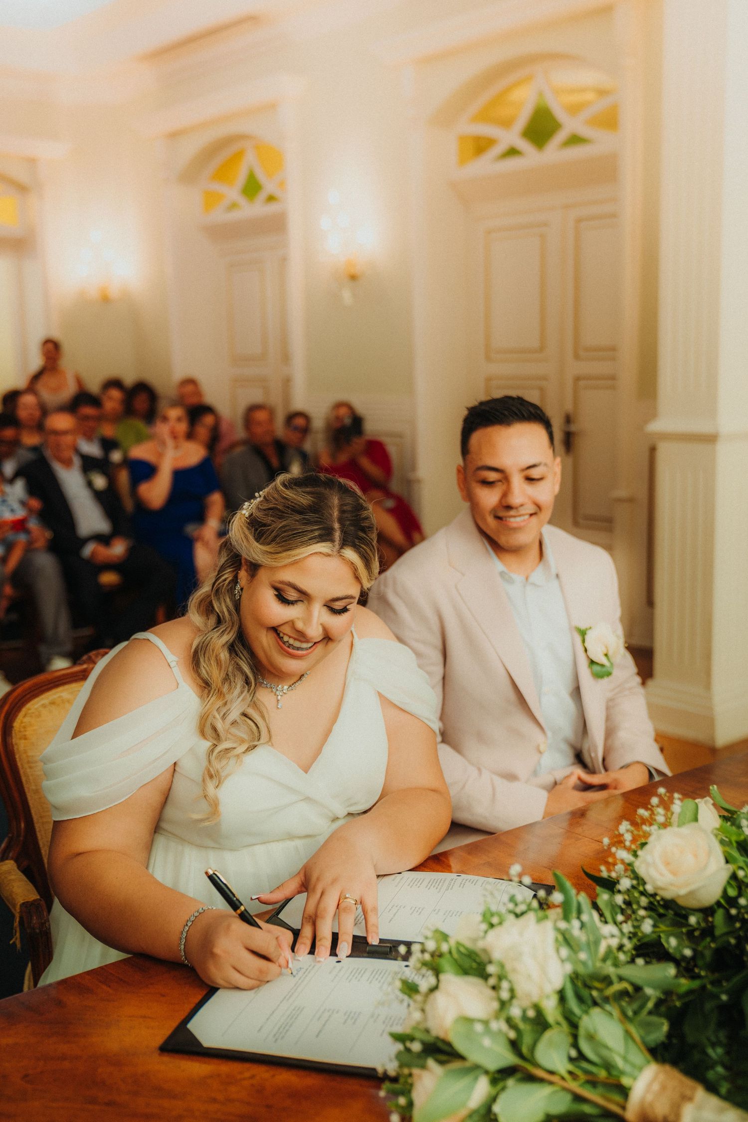 Wedding ceremony signing moment in elegant white room with guests watching.