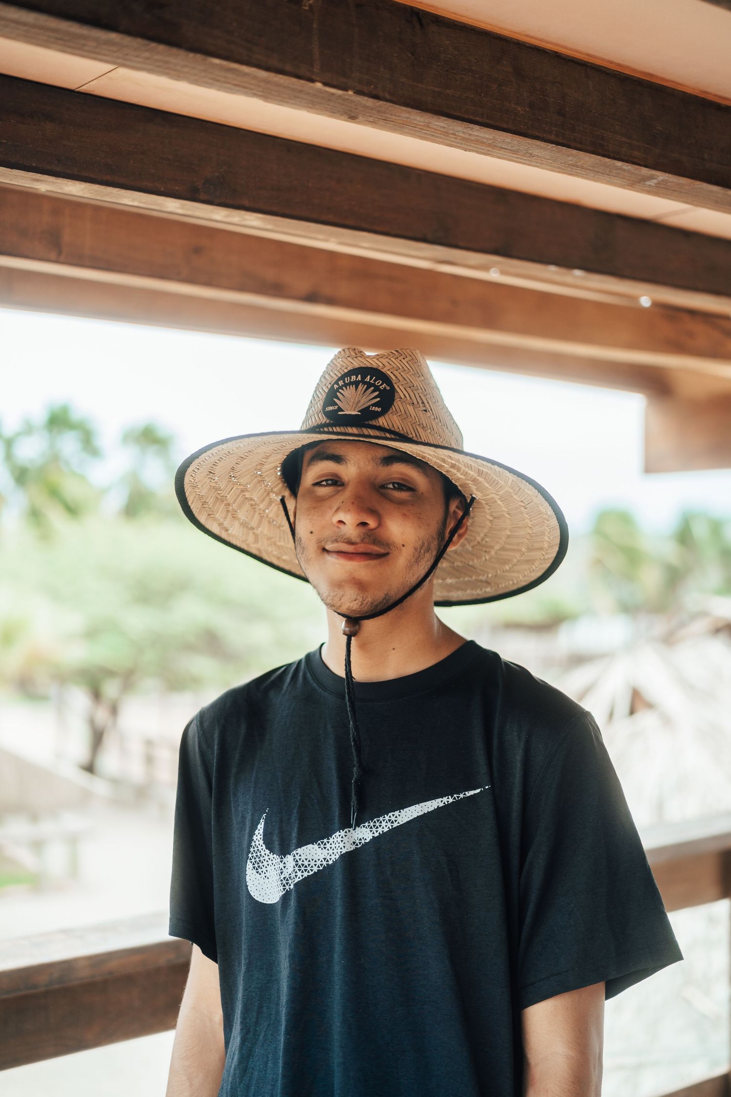 Person in black Nike shirt and straw hat posing under wooden shelter.