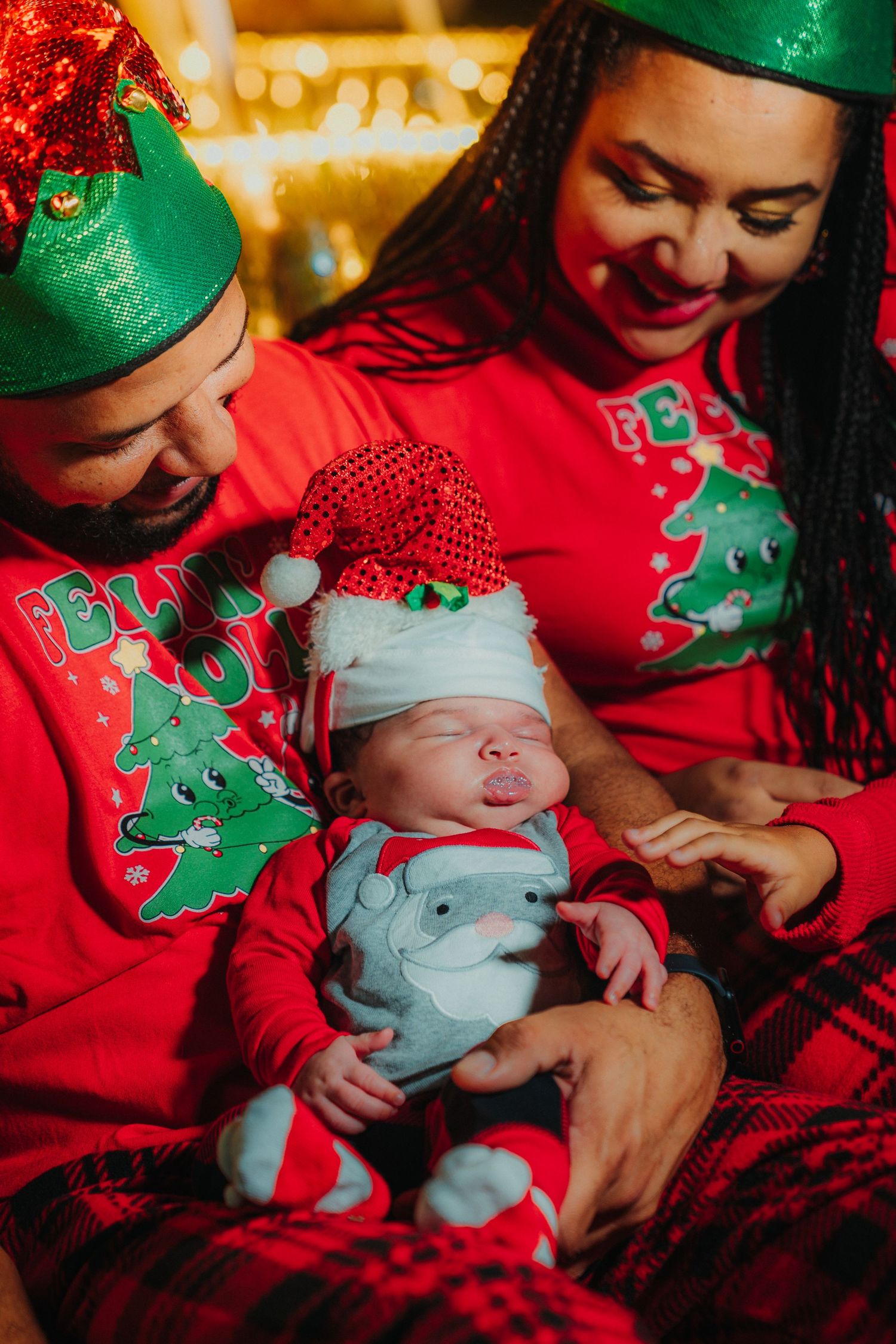 A family wearing festive red Christmas sweaters holds a baby in holiday attire against a warm glowing background.
