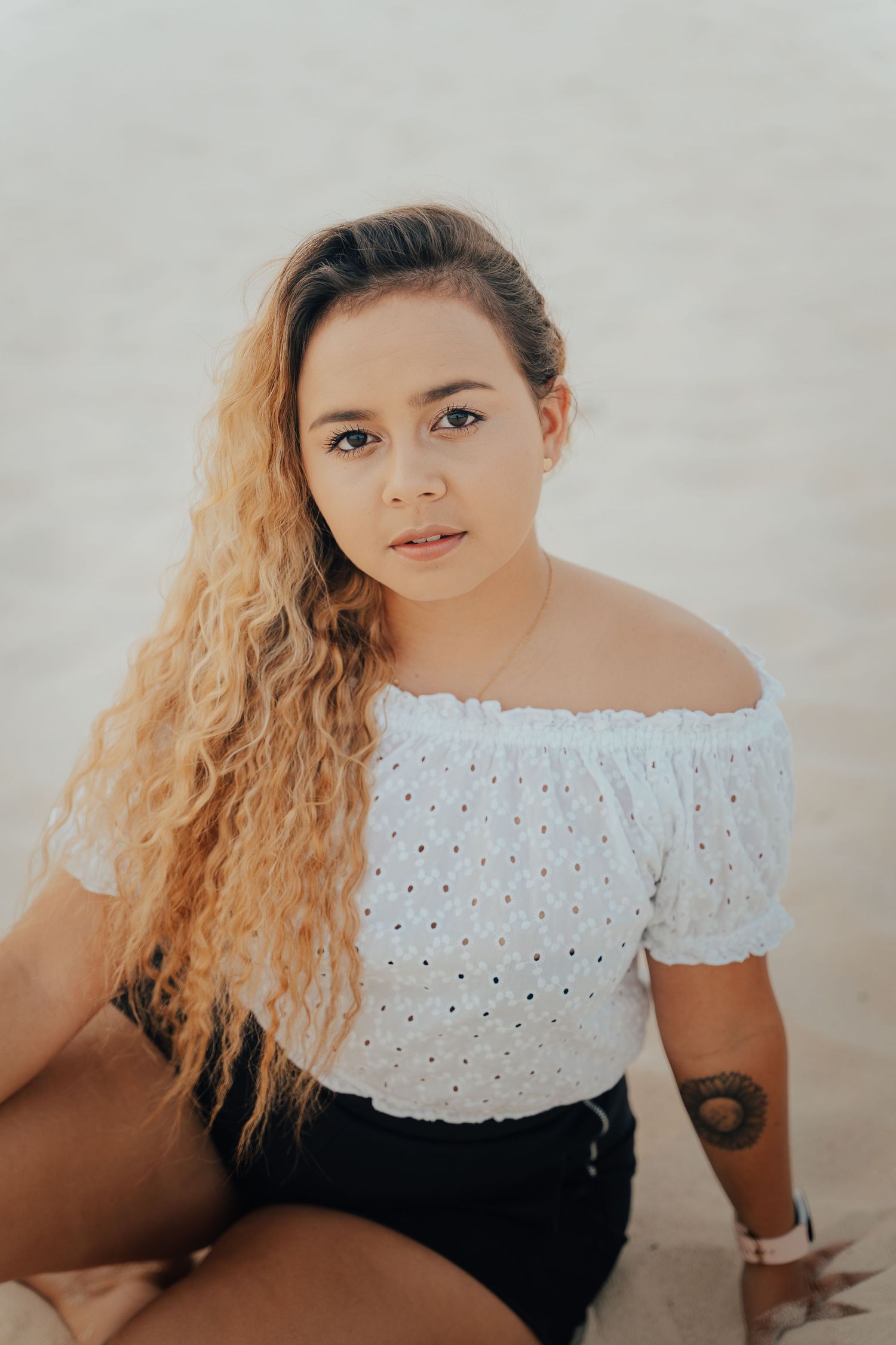 Woman in white eyelet off-shoulder top sitting on beach.