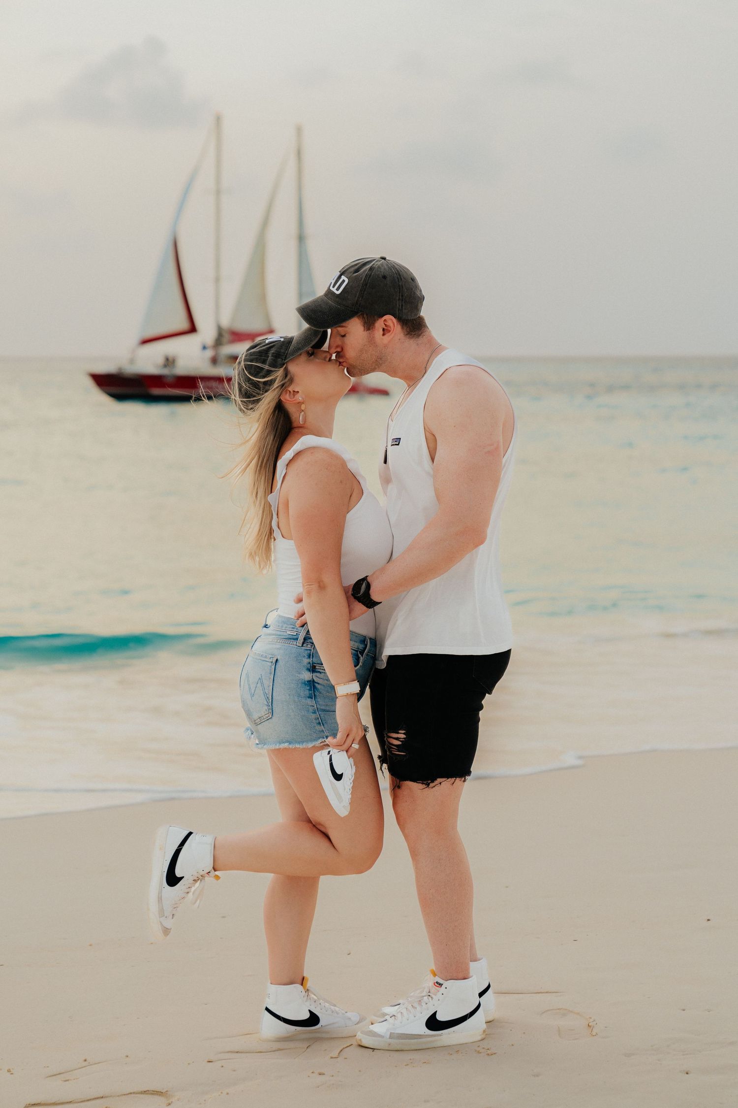 A romantic beachside moment with sailboats in the background during sunset.