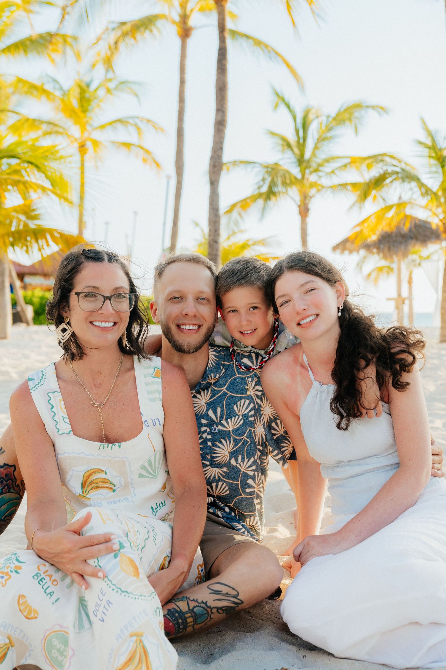 Beach family portrait with everyone wearing coordinated tropical and white outfits under palm trees.