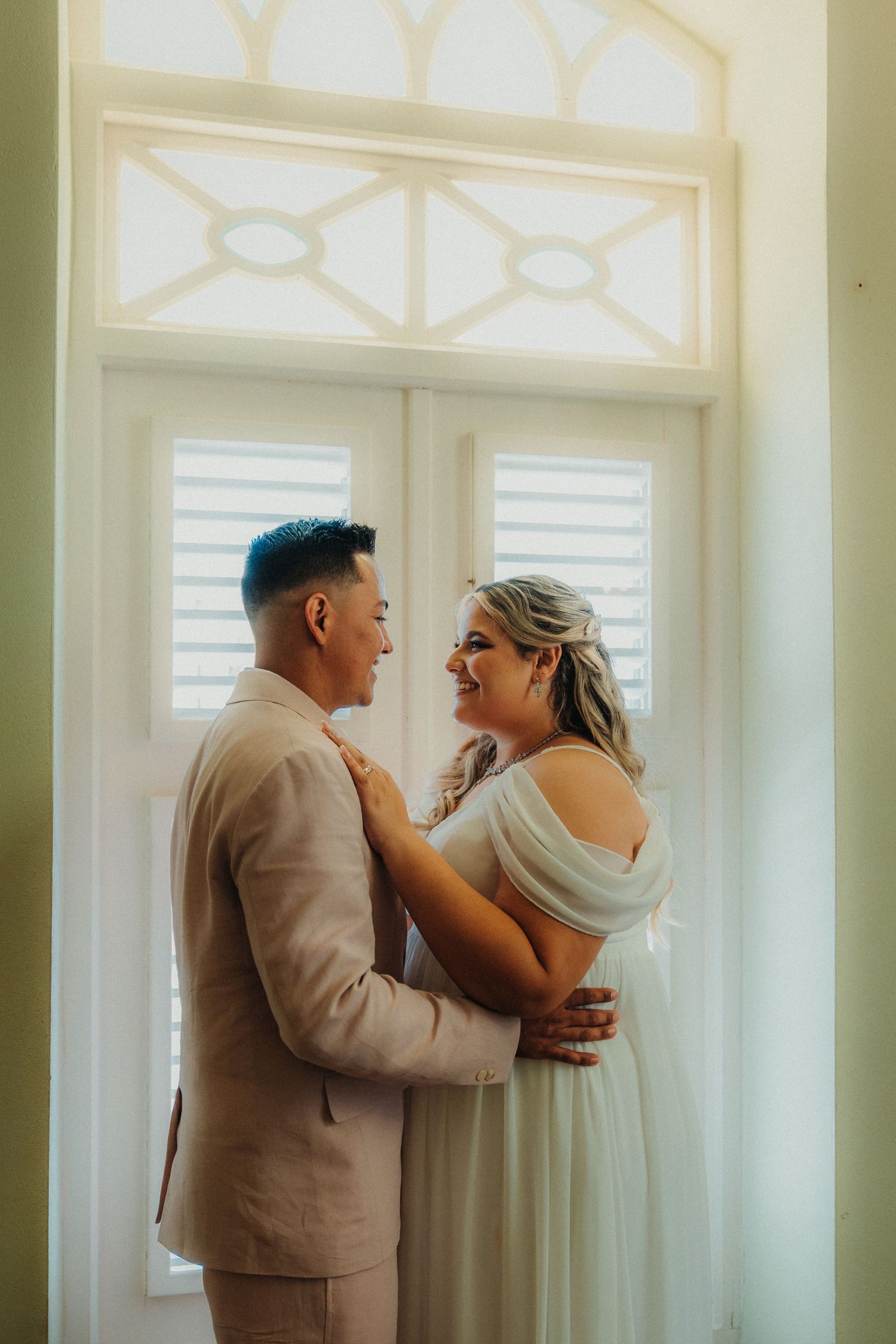 Couple in light colored formal wear share emotional moment by bright windows.