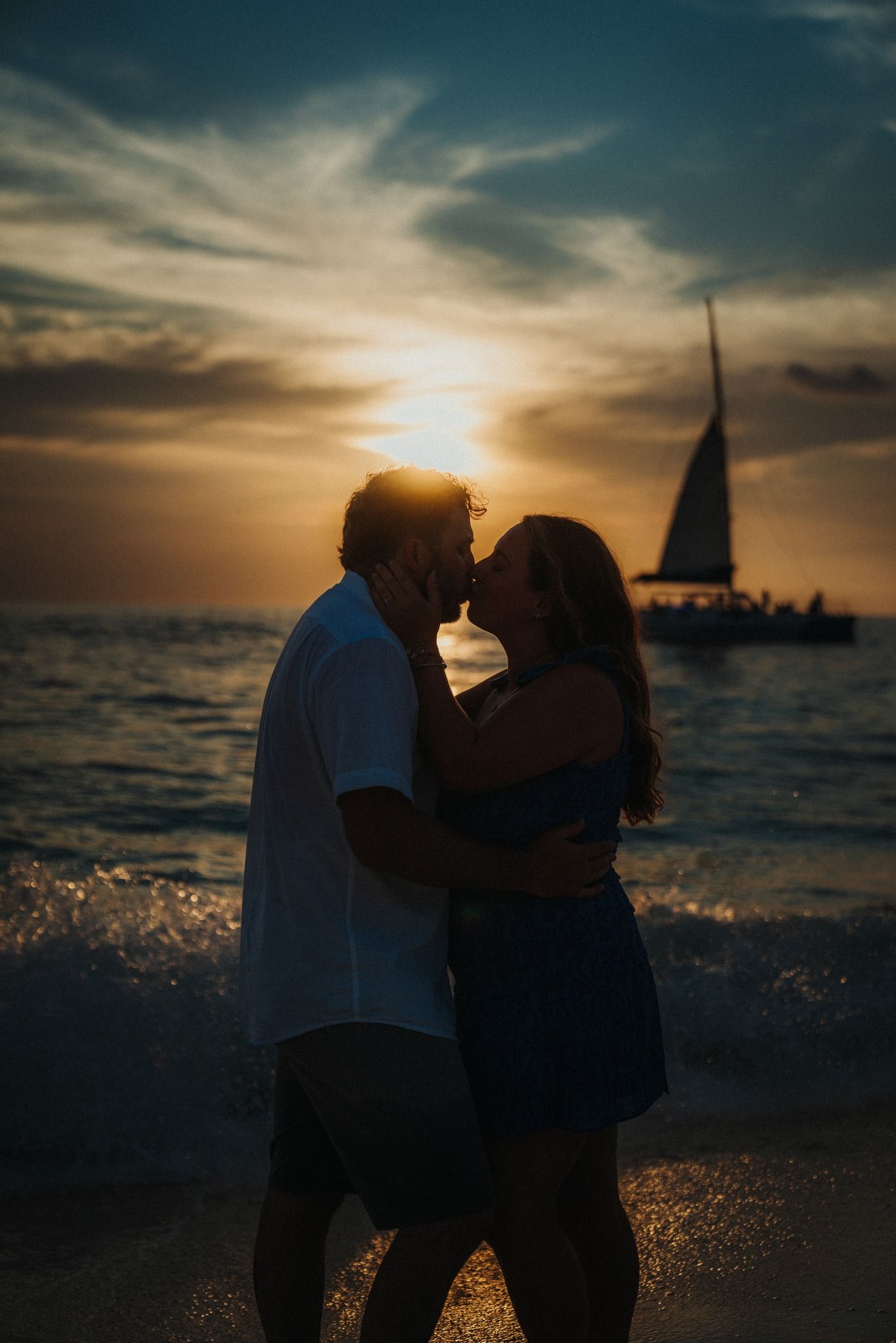 Silhouettes against a dramatic sunset sky over ocean waters with sailboats in the distance.