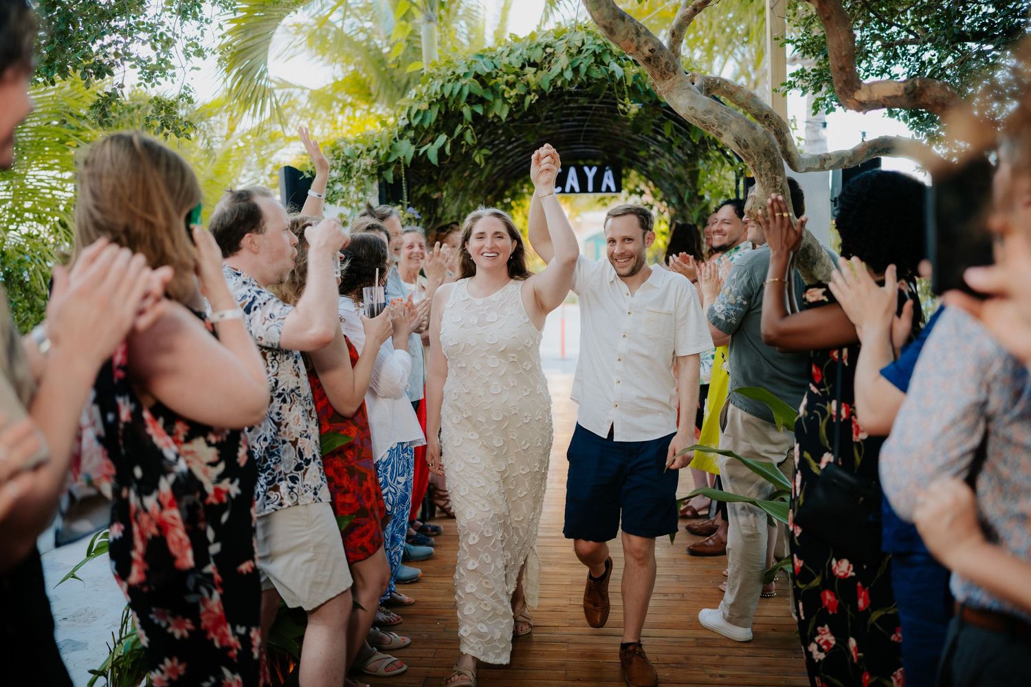 Joyful newlyweds raise hands walking through cheering guests at an outdoor tropical wedding celebration.