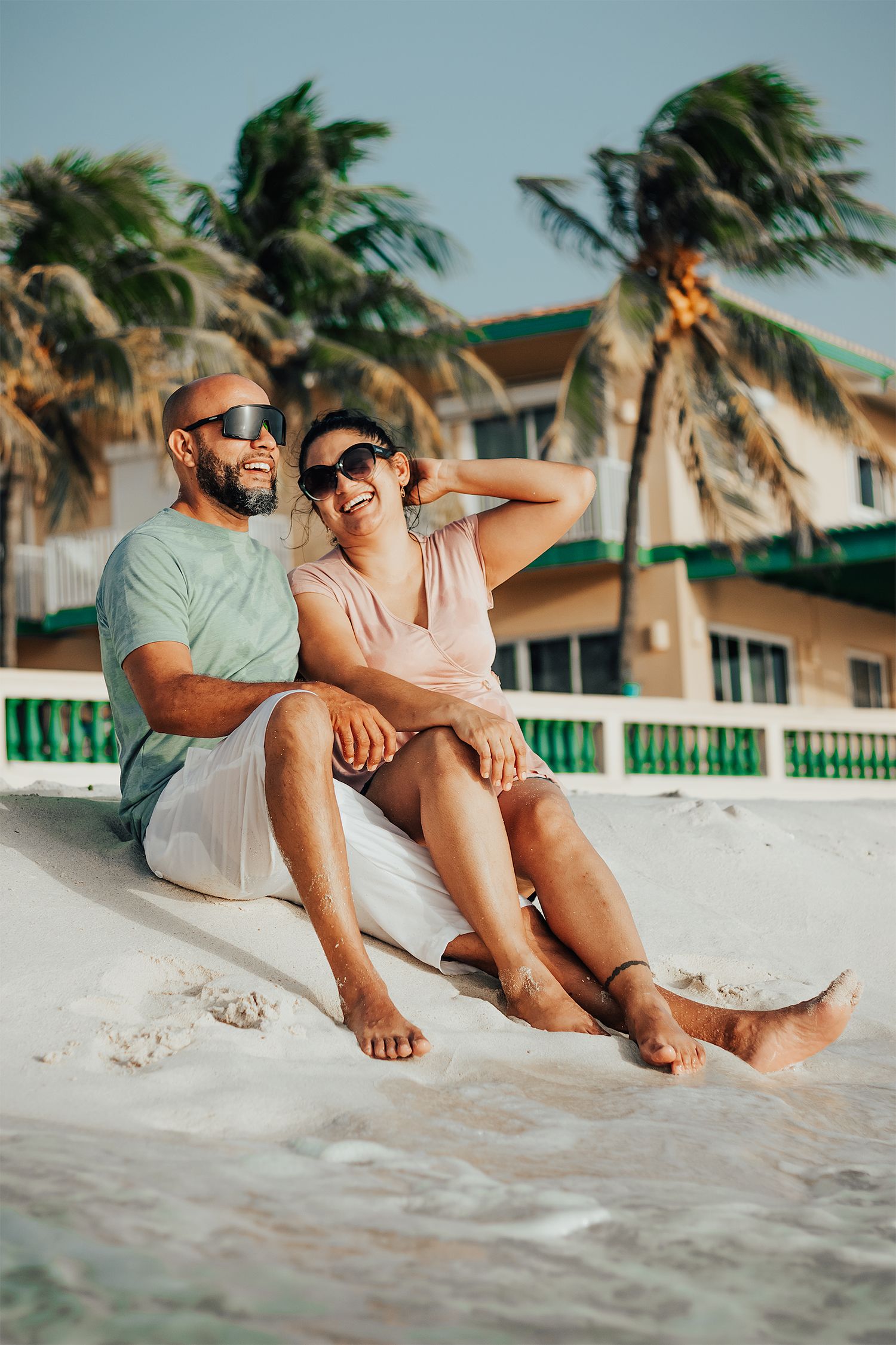 Couple relaxing on beach sand with palm trees swaying in background.