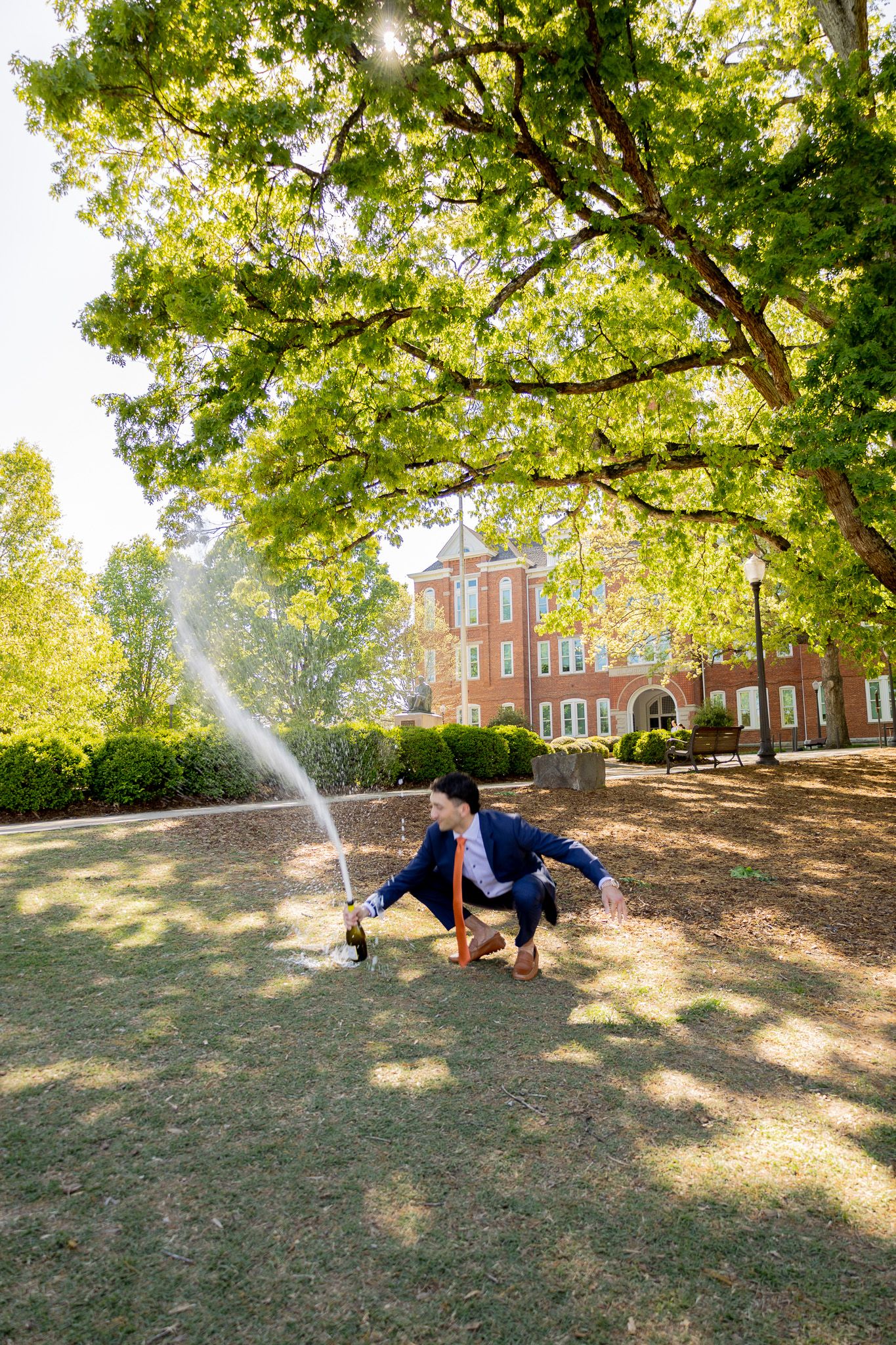 Graduate in cap and gown pops champagne bottle on college campus lawn beneath lush green trees.