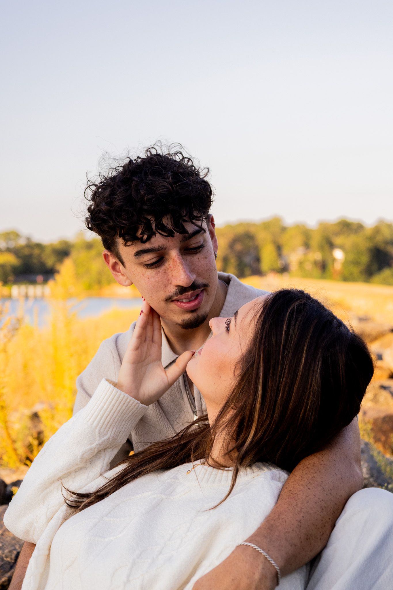 A young couple shares a tender moment outdoors, with the woman gently touching the man's face against a golden autumn backdrop.