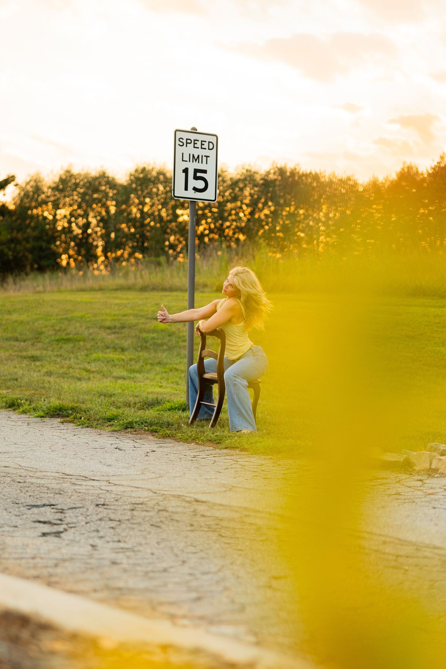 Blonde woman in yellow top leans against a speed limit 15 sign with a guitar at golden hour sunset.