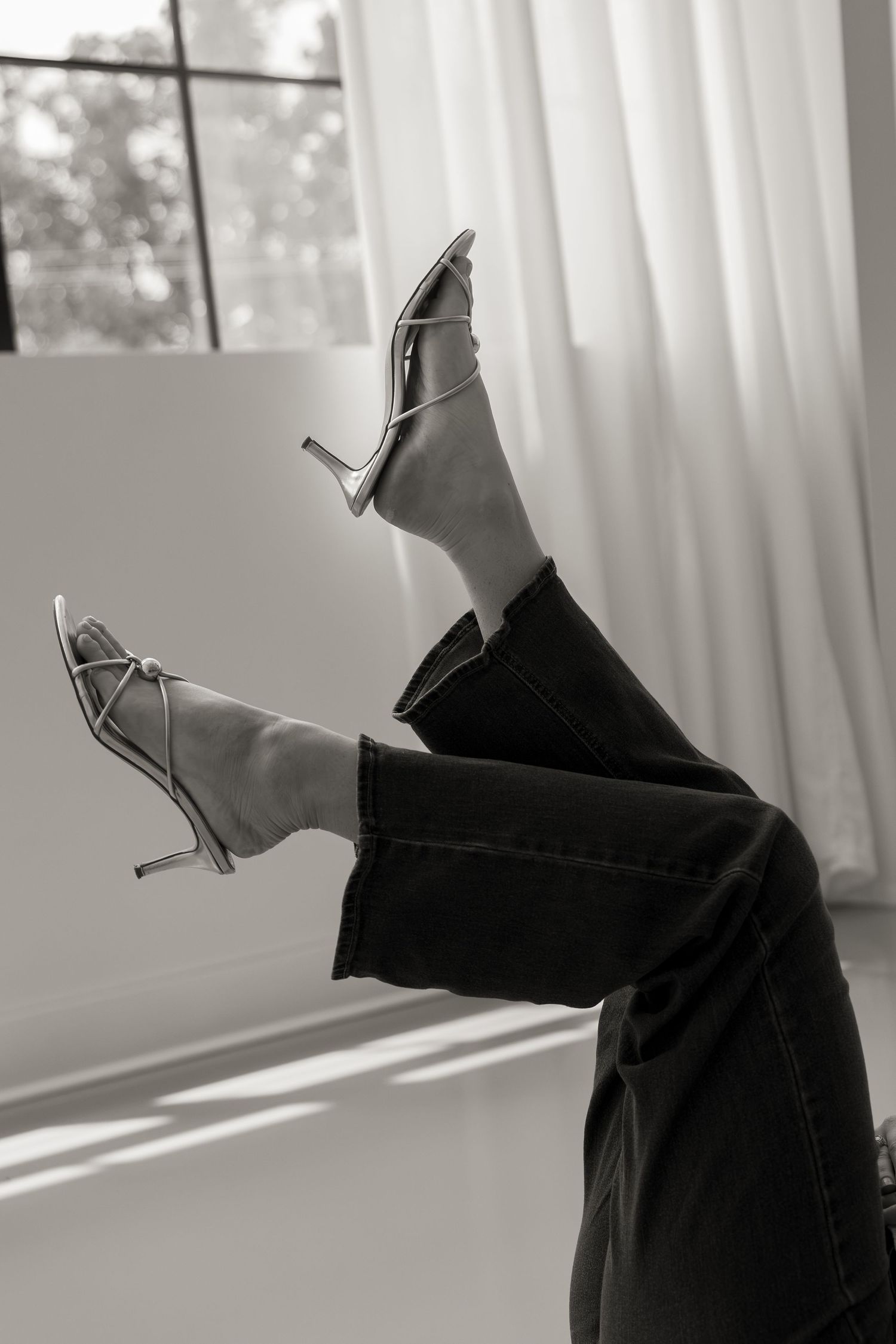 Black and white photo of legs raised in the air wearing strappy stiletto heels and dark jeans near a window.
