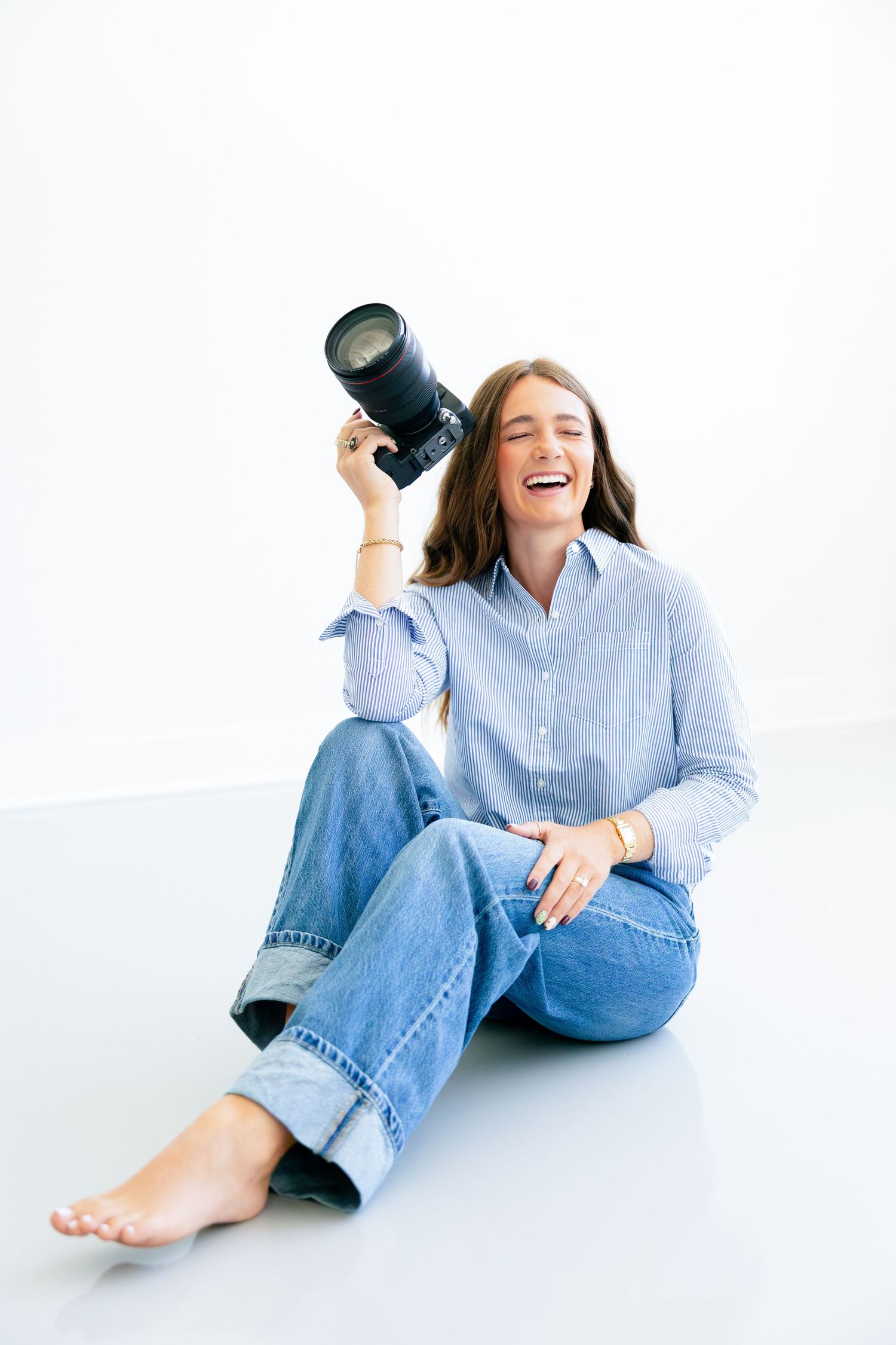 A laughing woman sits on a white floor holding a DSLR camera, wearing a blue shirt and jeans.