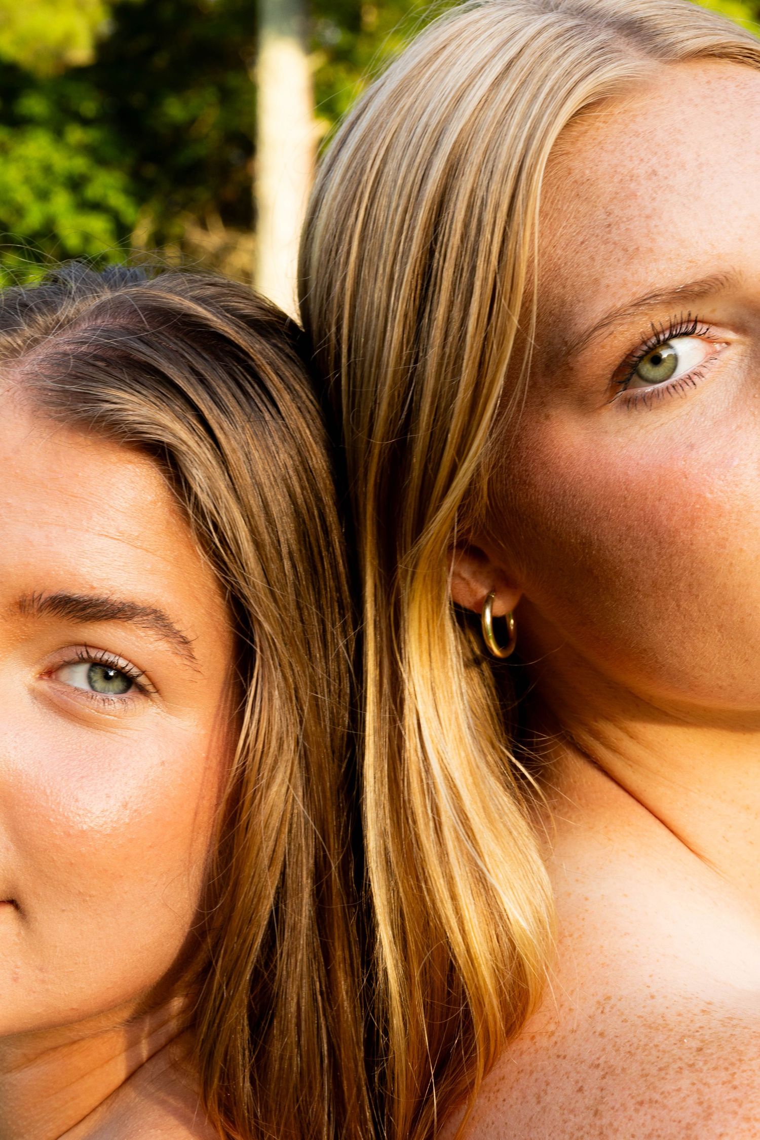 Two young women with blonde and brunette hair pose back-to-back outdoors, both with green eyes and freckles.