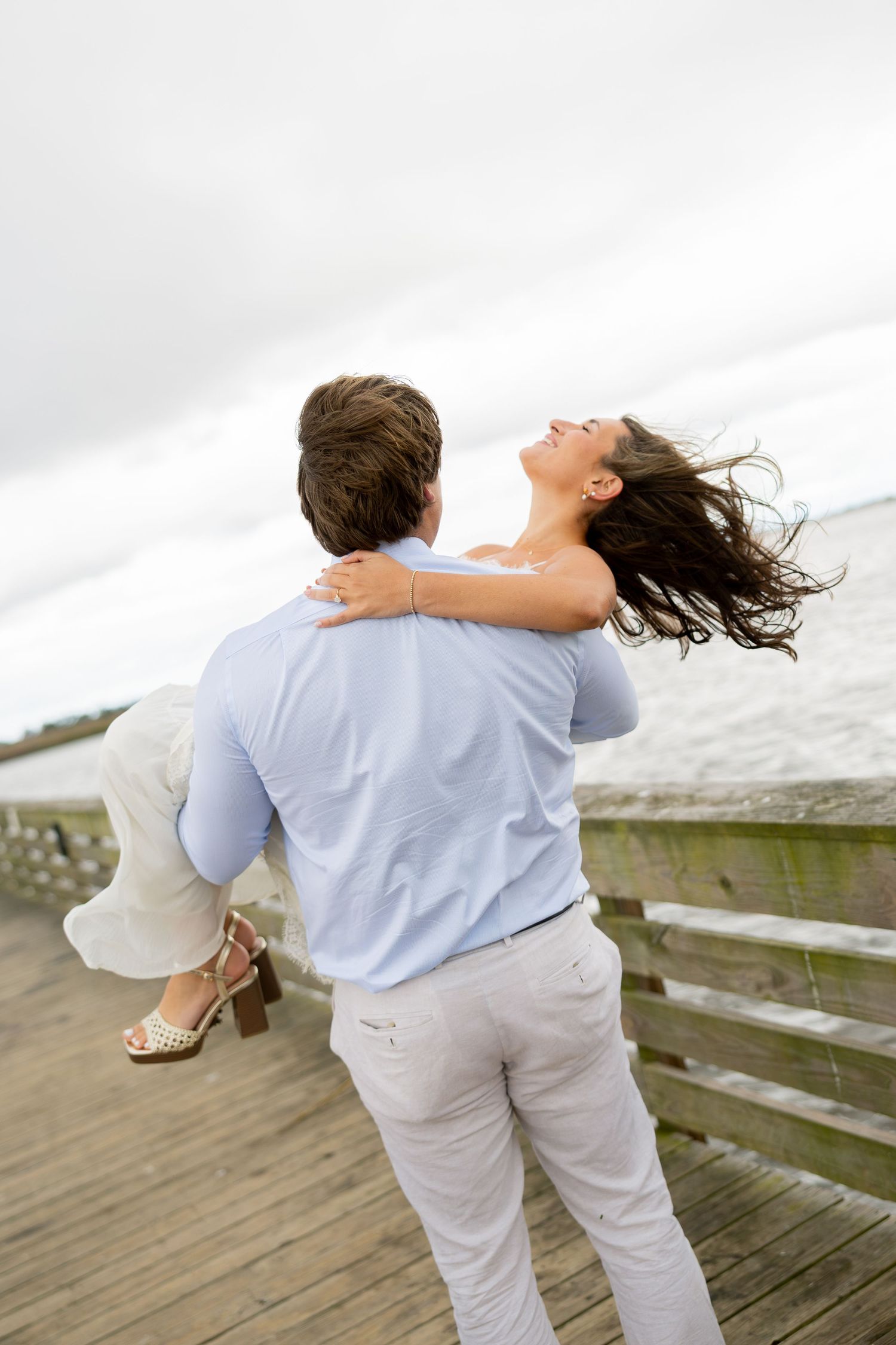A man in a light blue shirt carries a laughing woman with windblown hair in his arms on a wooden pier overlooking the water.
