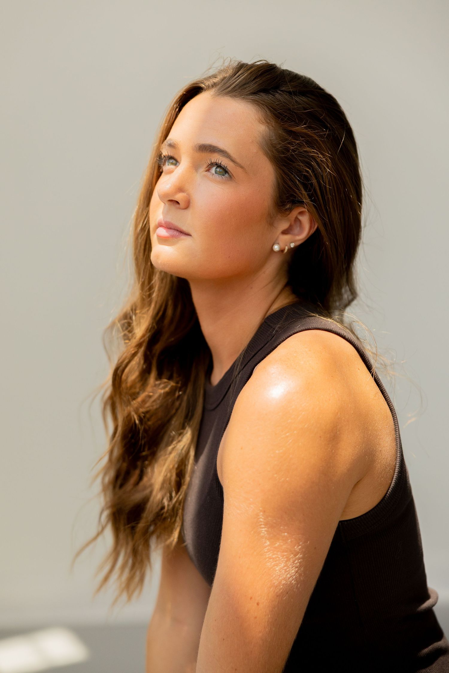 Young woman with long brown hair wearing a black sleeveless top, gazing upward in natural sunlight.