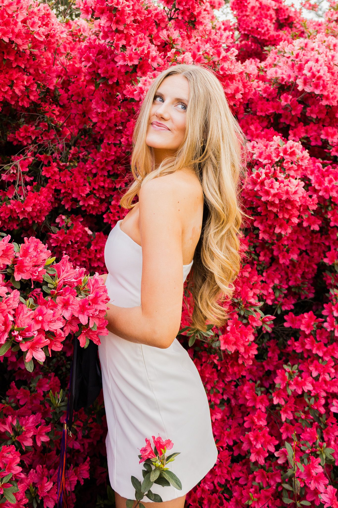 Young blonde woman in white dress smiling among vibrant pink azalea flowers in full bloom.