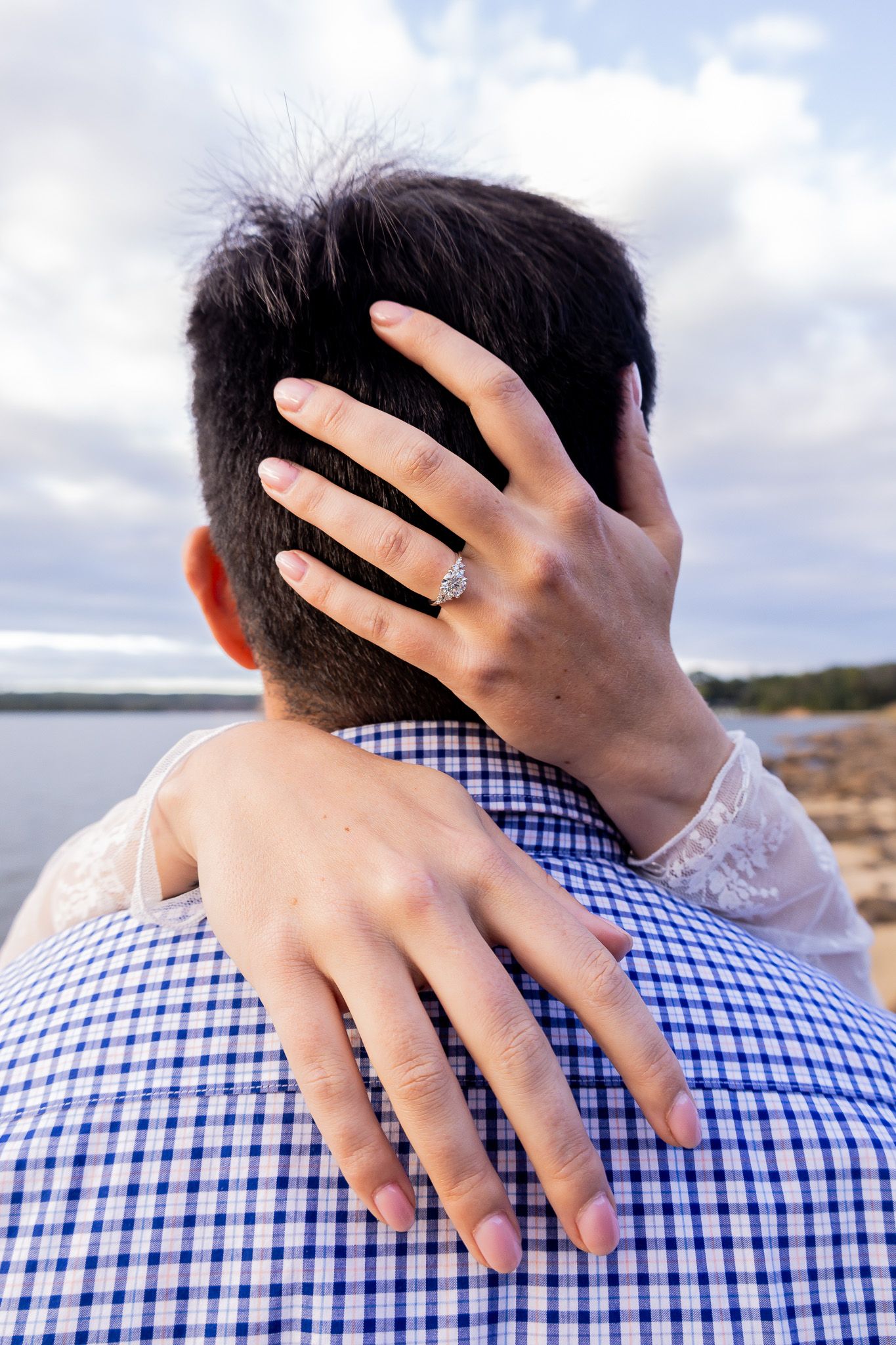Close-up of engagement ring on woman's hand embracing man in blue plaid shirt by the water.