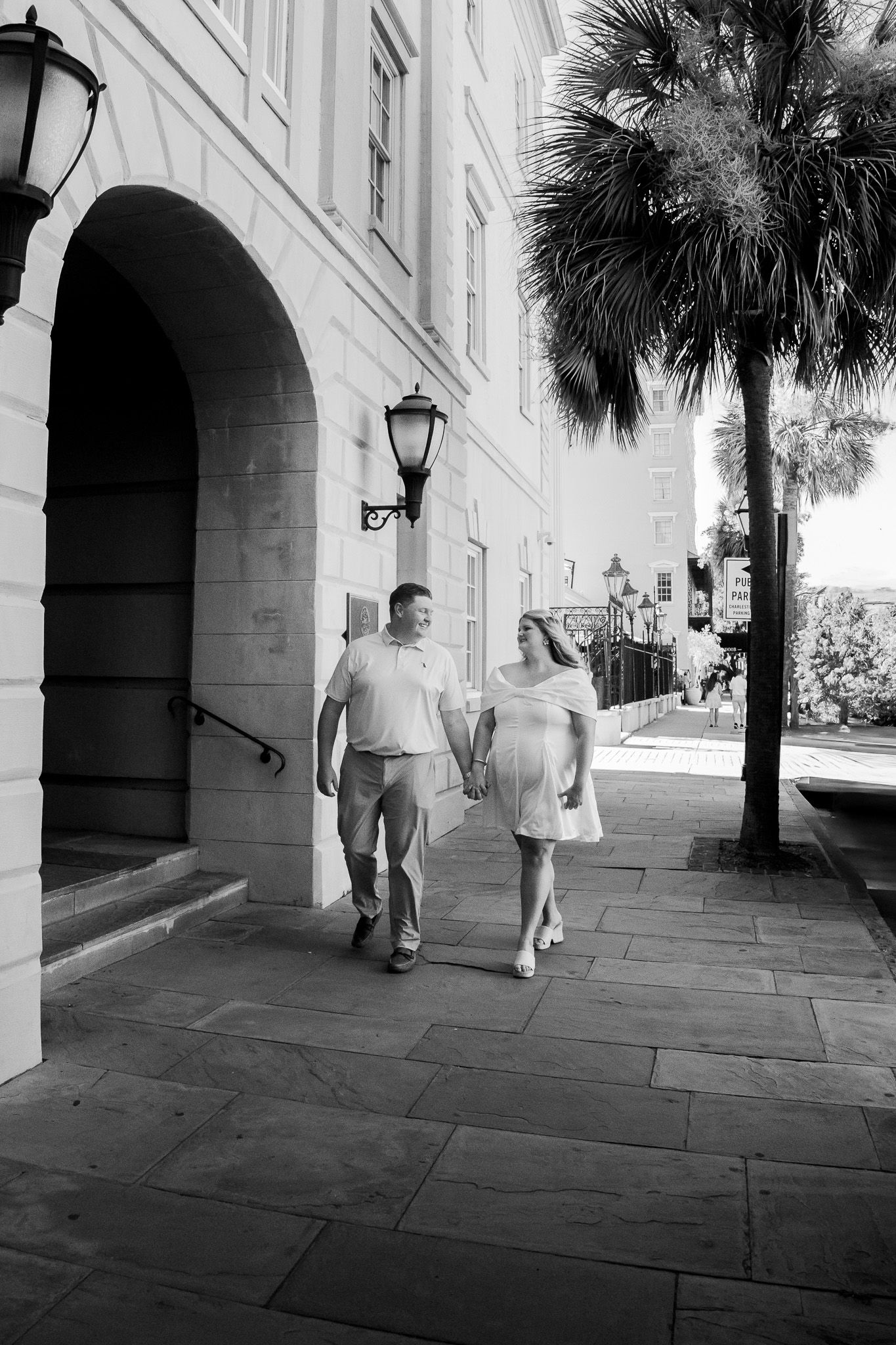 Black and white photo of a couple holding hands walking along a historic building sidewalk with palm trees.