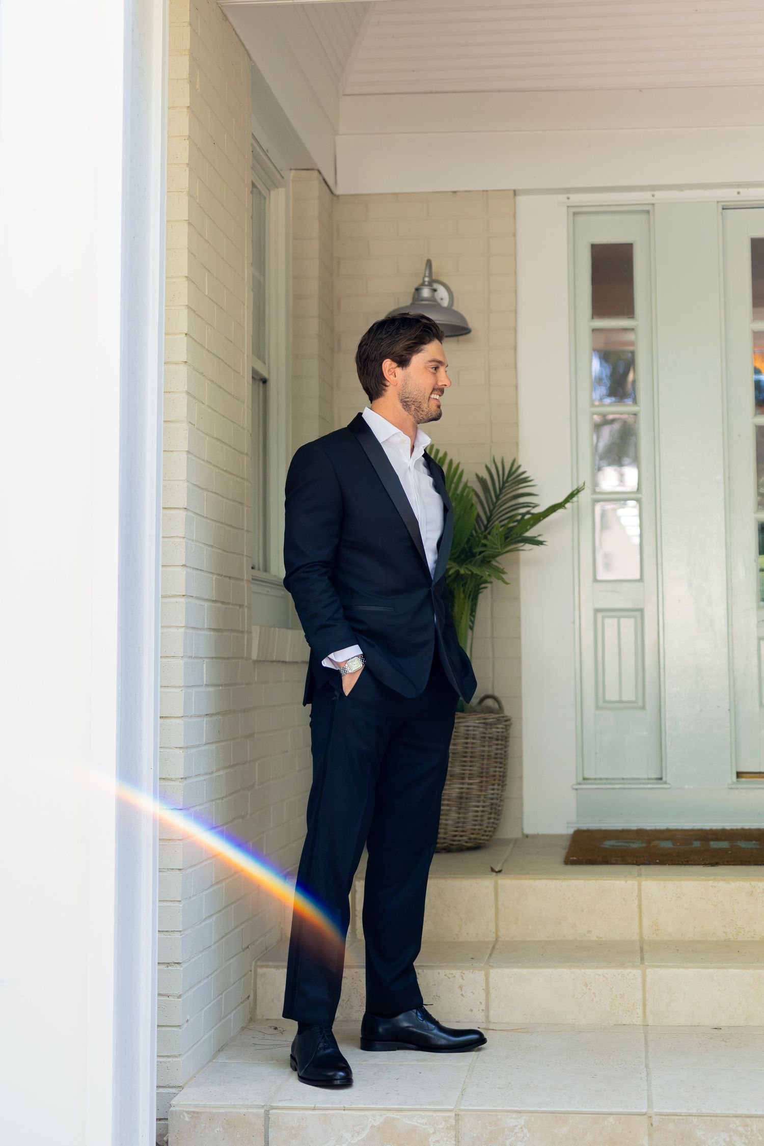 Man in dark suit standing on front porch steps, hands in pockets, looking to the side near a green door.