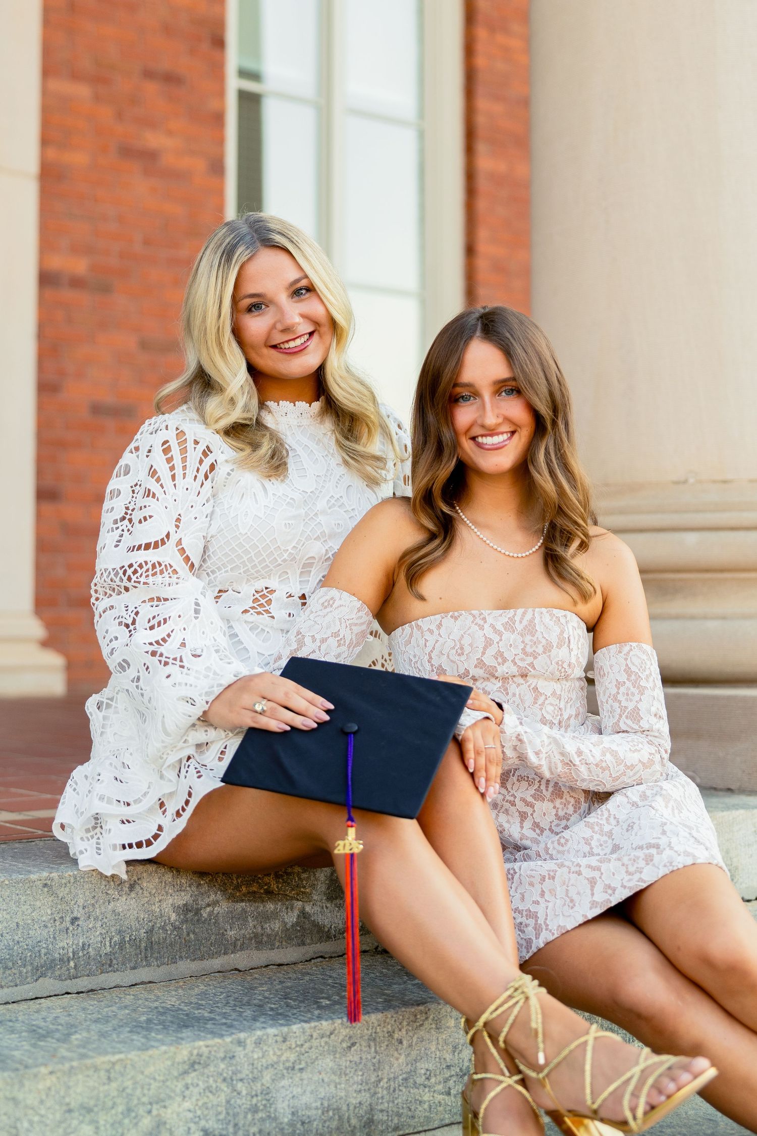 Two smiling women in white lace dresses pose on steps holding a graduation cap during a college graduation celebration.