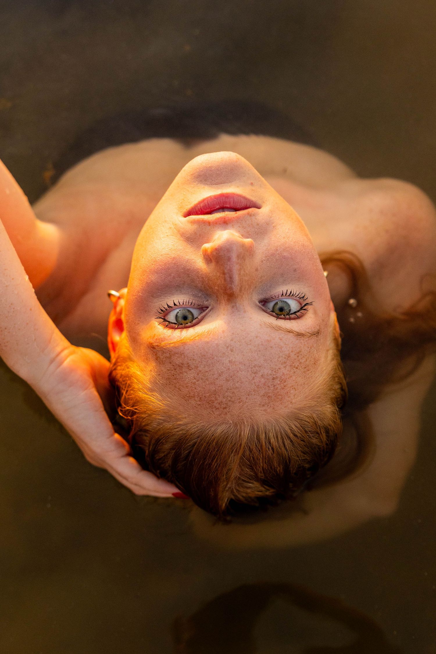 A freckled redhead woman with blue eyes floats on her back in dark water, looking up at the camera.