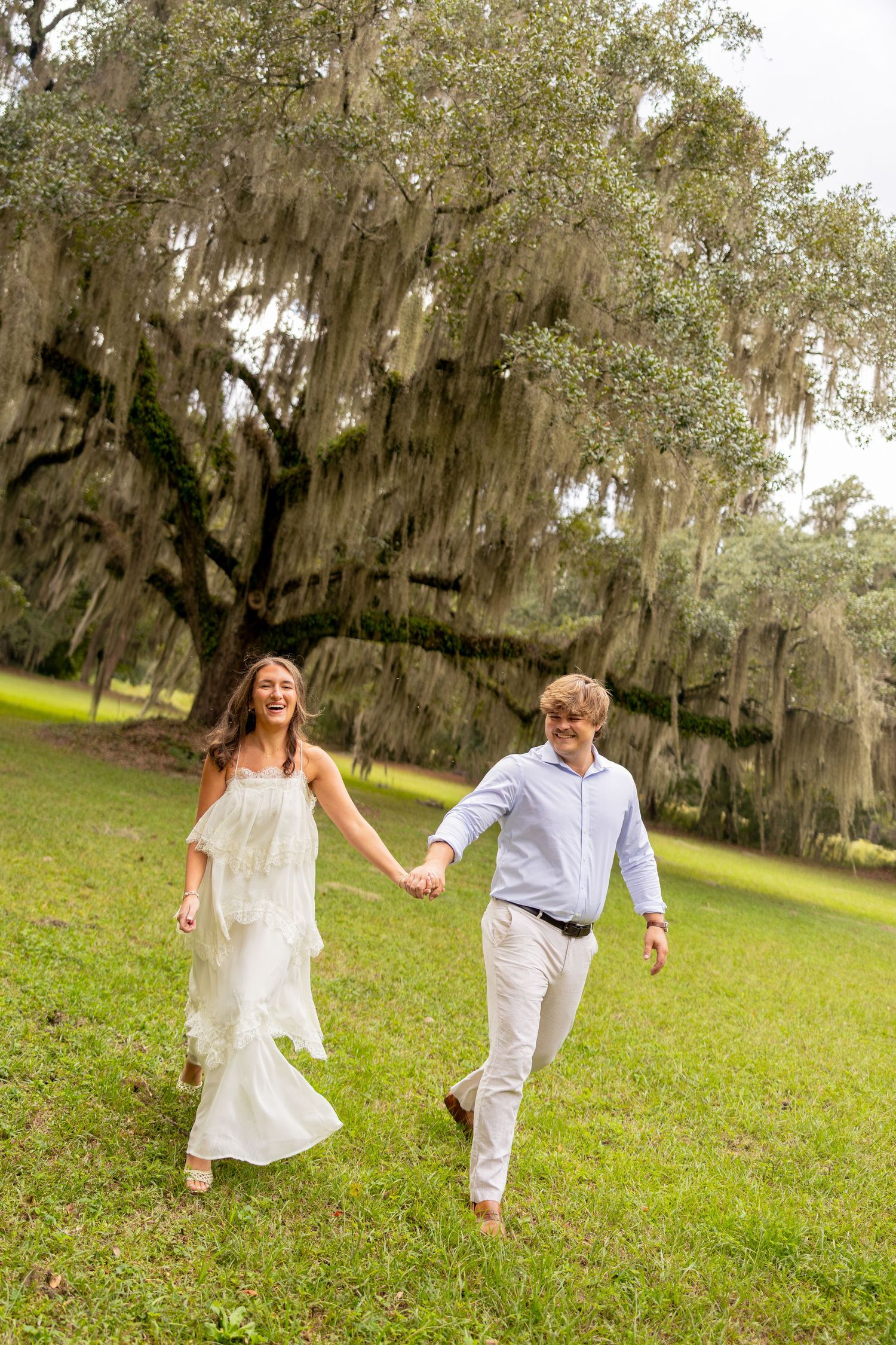 Smiling couple holding hands and walking barefoot across a green lawn beneath a large live oak draped in Spanish moss.