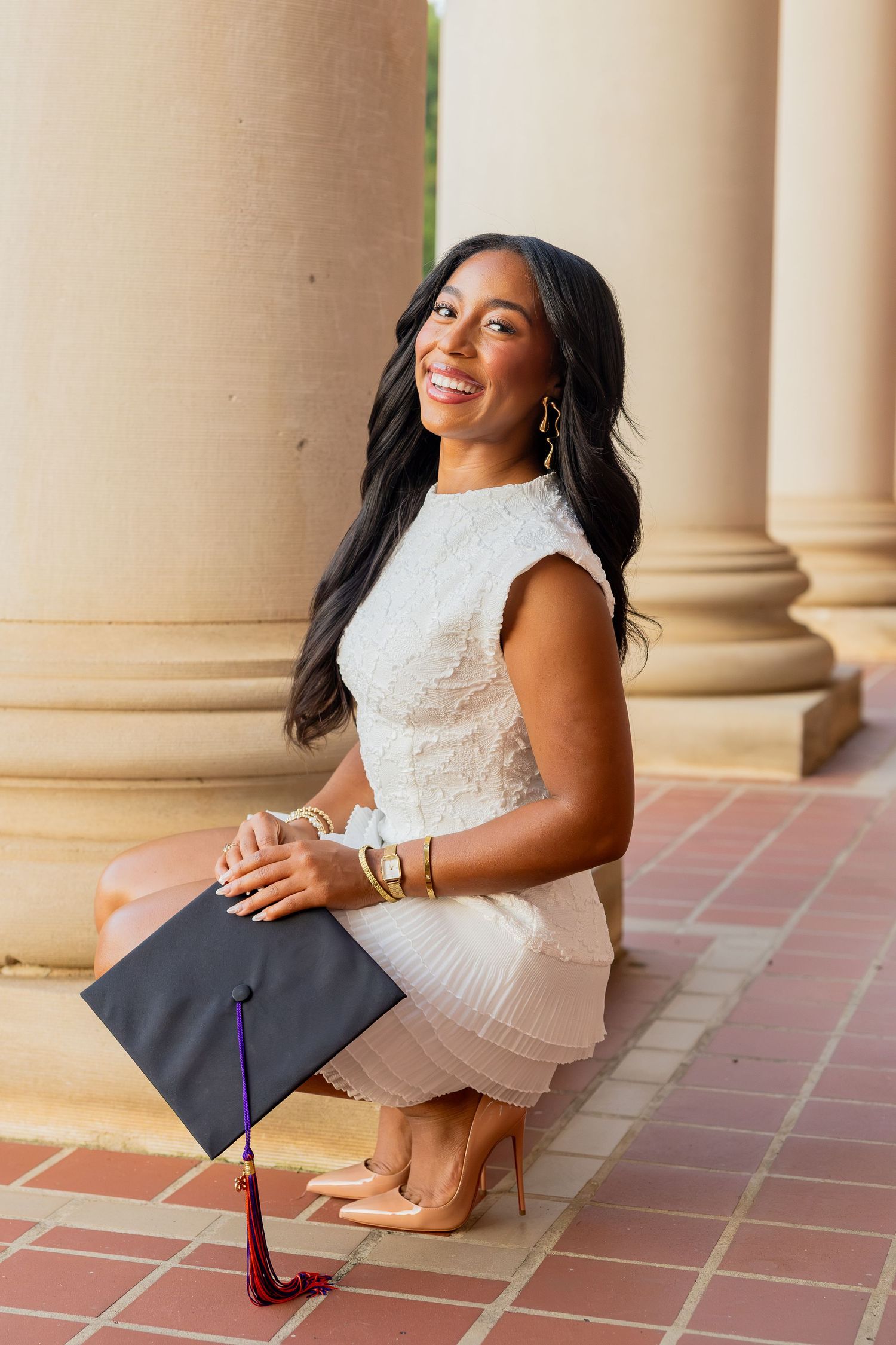 Smiling young woman in white dress holding graduation cap while sitting on brick steps near columns.