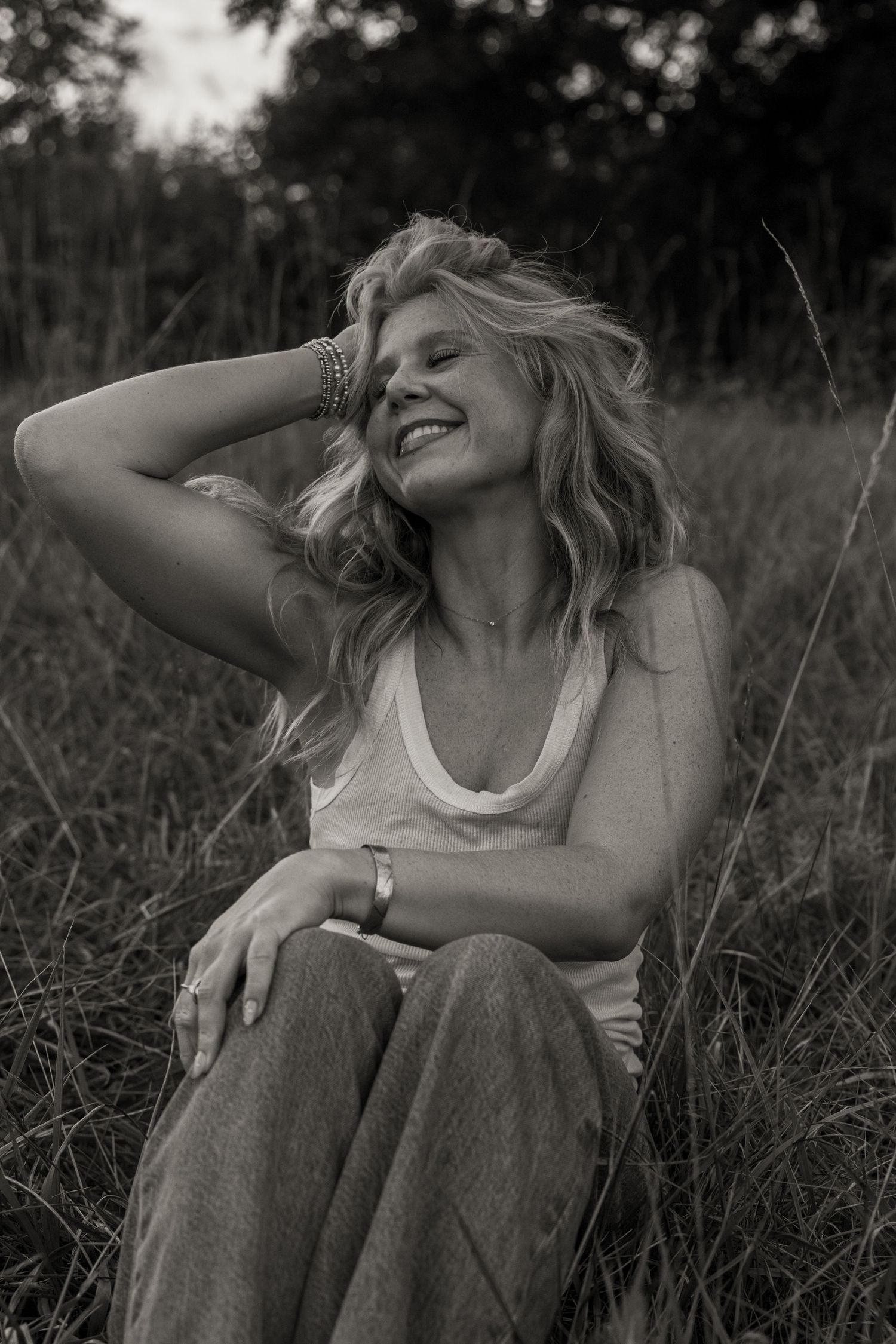 Black and white photo of a smiling woman sitting in a grassy field, hand in hair, wearing a tank top and wide-leg jeans.
