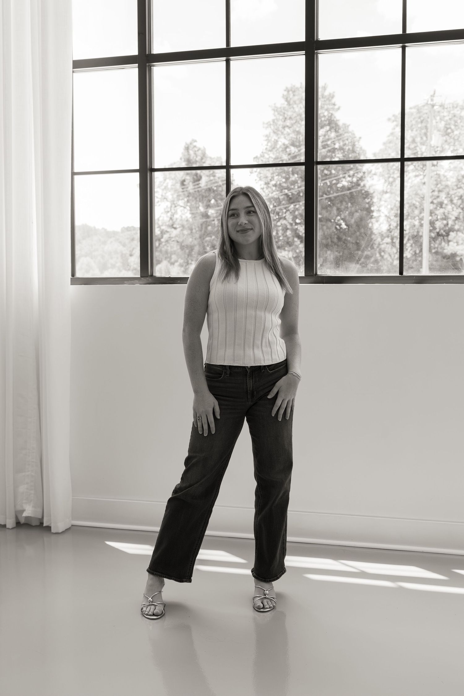 Black and white photo of a young woman in a ribbed tank top and wide-leg jeans posing in front of a large window.